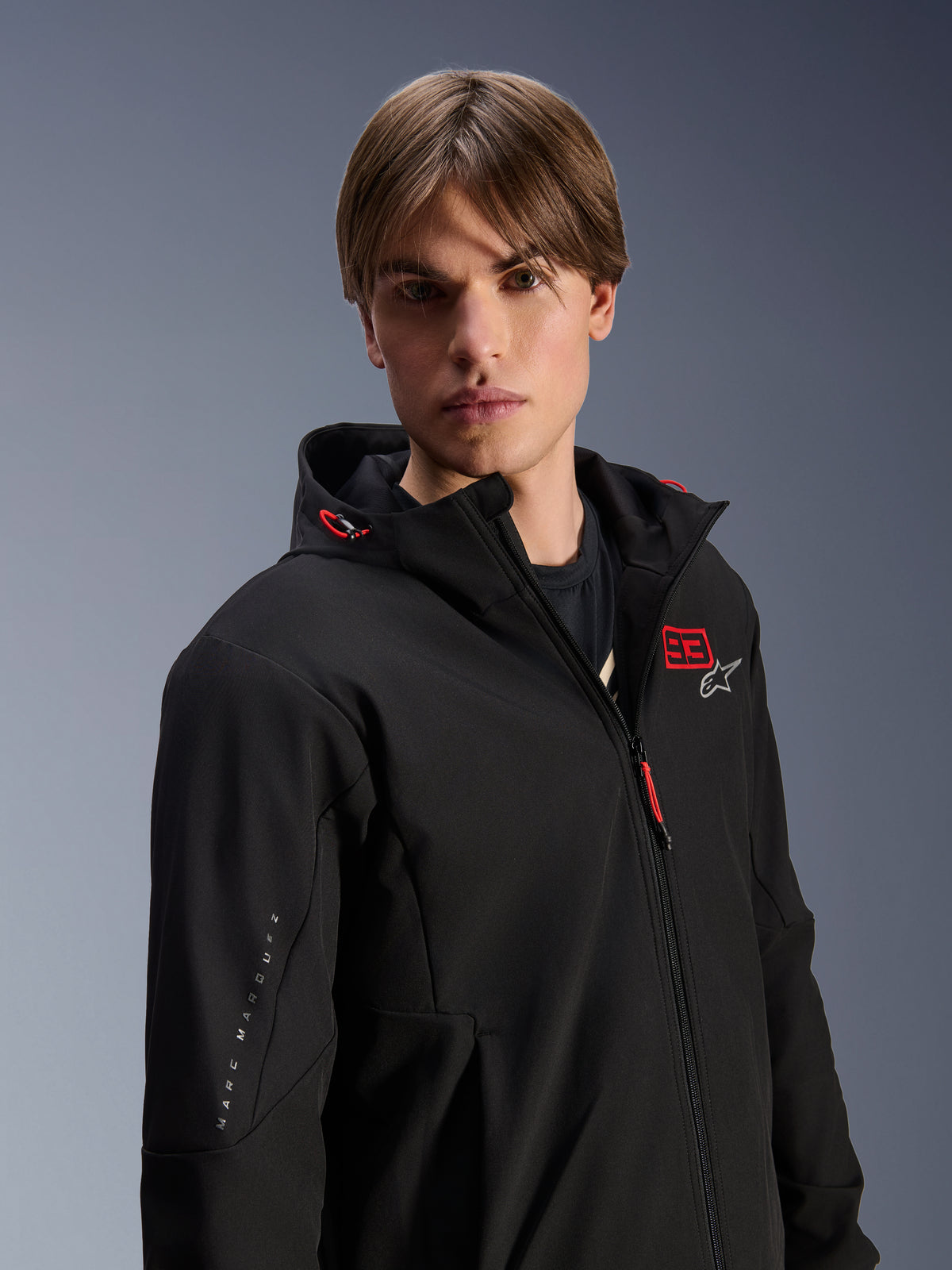 Mannelijk model draagt Alpinestars MM93 Radium Tech Hoodie, zwarte urban motorjas, voorzien van rood '93' en wit sterlogo op de borst, 'Marc Marquez' branding op de mouw, technische winddichte shell voor stedelijk rijden.