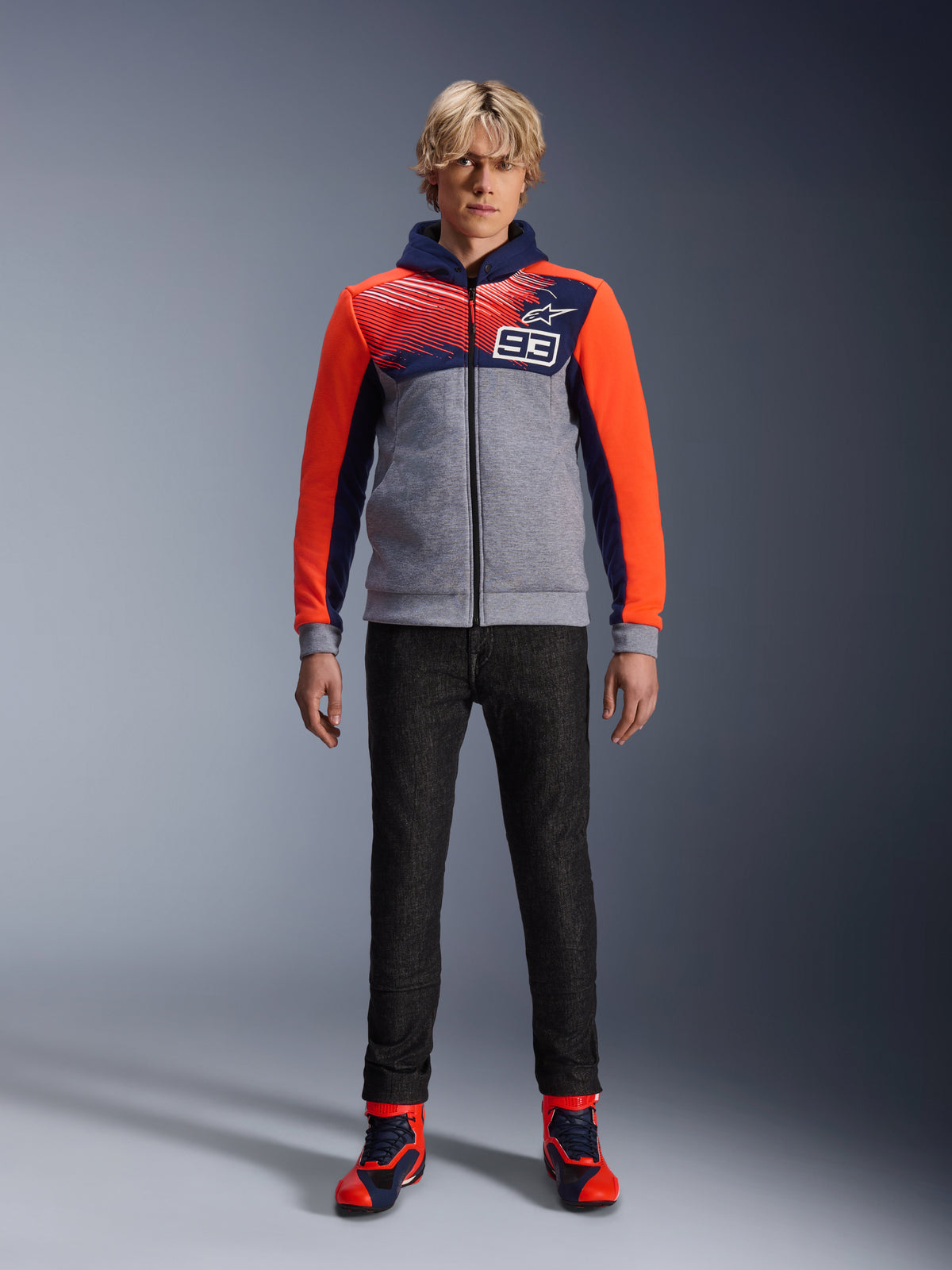 Alpinestars MM93 Chrome V2 Sport Hoodie, motorhoodie, Blue Navy Gray Melange en Red Fluo, gedragen met een donkere motorbroek en sneakers met rode accenten, voorzien van een doorlopende ritssluiting en MM93-logo, casual urban motorsport-geïnspireerd ontwerp