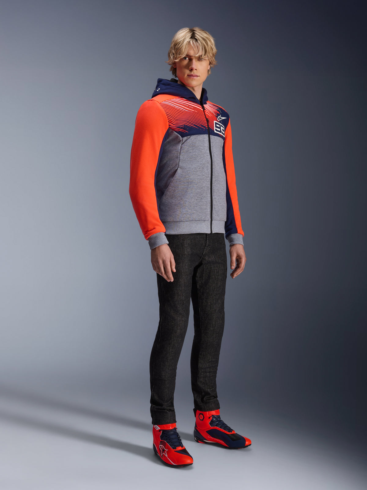 Alpinestars MM93 Chrome V2 Sport Hoodie, urban motorhoodie, grijs gemêleerde body met donkerblauwe borst en felrode fluo mouwen, voorzien van het 93-logo op de borst, gedragen door een model met slim-fit zwarte broek en bijpassende rode en blauwe hoge motorschoenen, moderne atletische pasvorm voor rijden op de weg