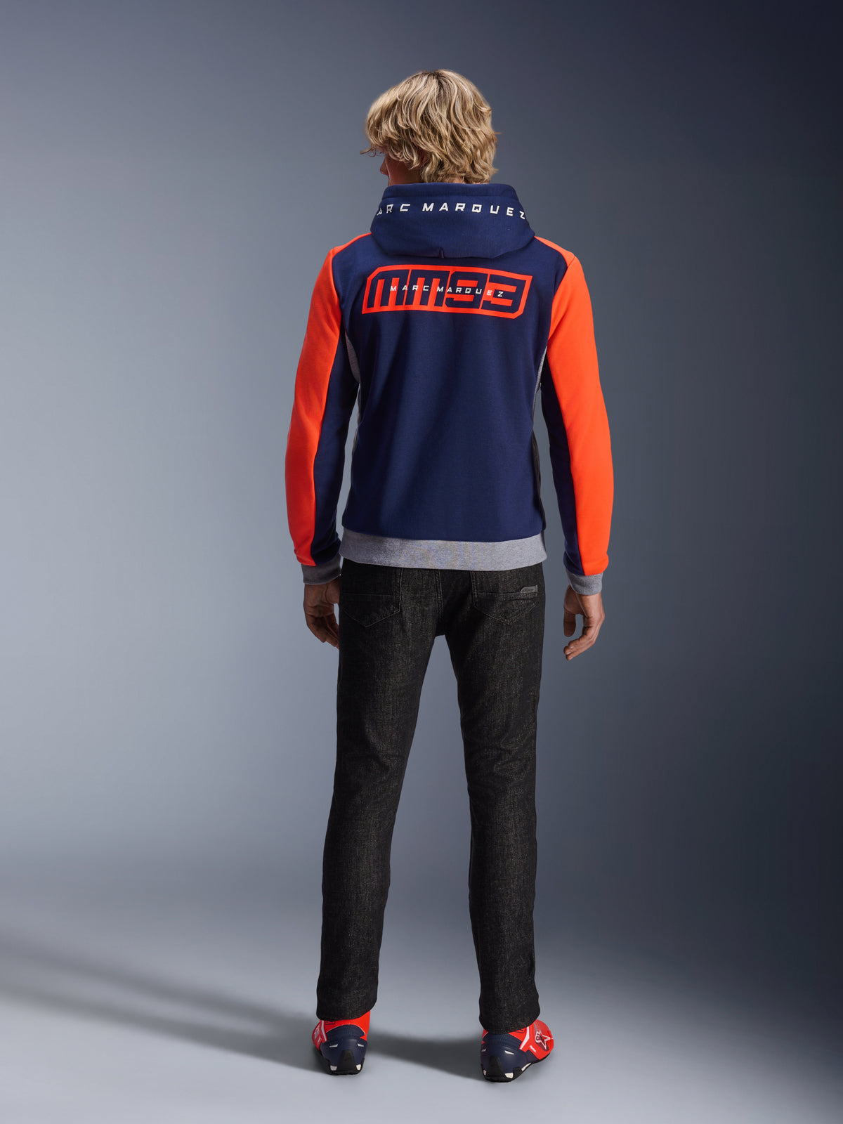 Model van achteren die de Alpinestars MM93 Chrome V2 Sport Hoodie draagt, donkerblauw met felrode fluo mouwen en grijs gemêleerde details, grote MM93- en Marc Marquez-logo's op de rug en capuchon, casual motorhoodie gecombineerd met een zwarte broek