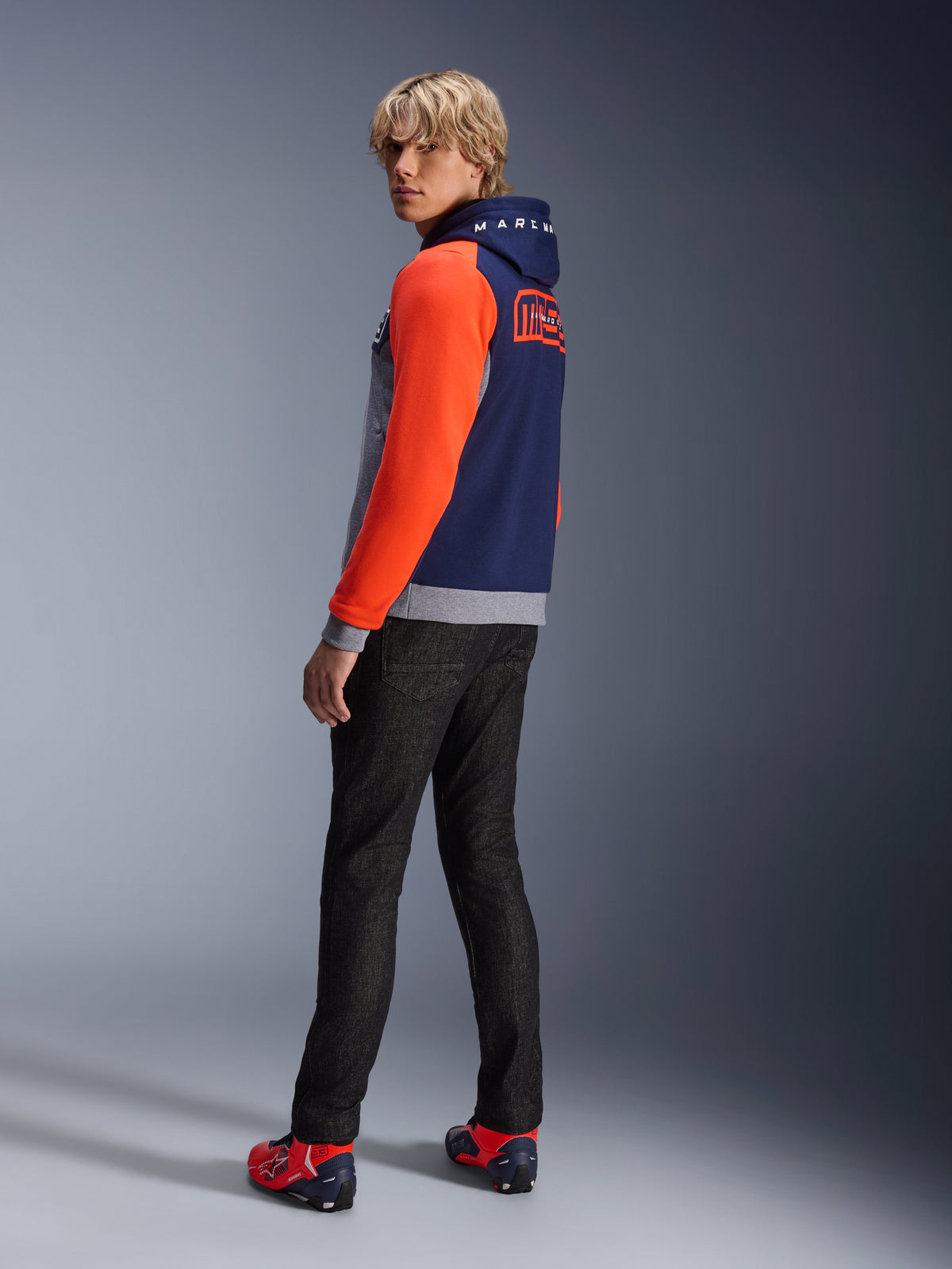 Alpinestars MM93 Chrome V2 Sport Hoodie, Blue Navy Gray Mel Red Fluo, model draagt hoodie met felrode mouwen en MM93 Marc Marquez branding op de rug, gecombineerd met een donkere jeans en rode motorschoenen, casual stedelijke motorkleding