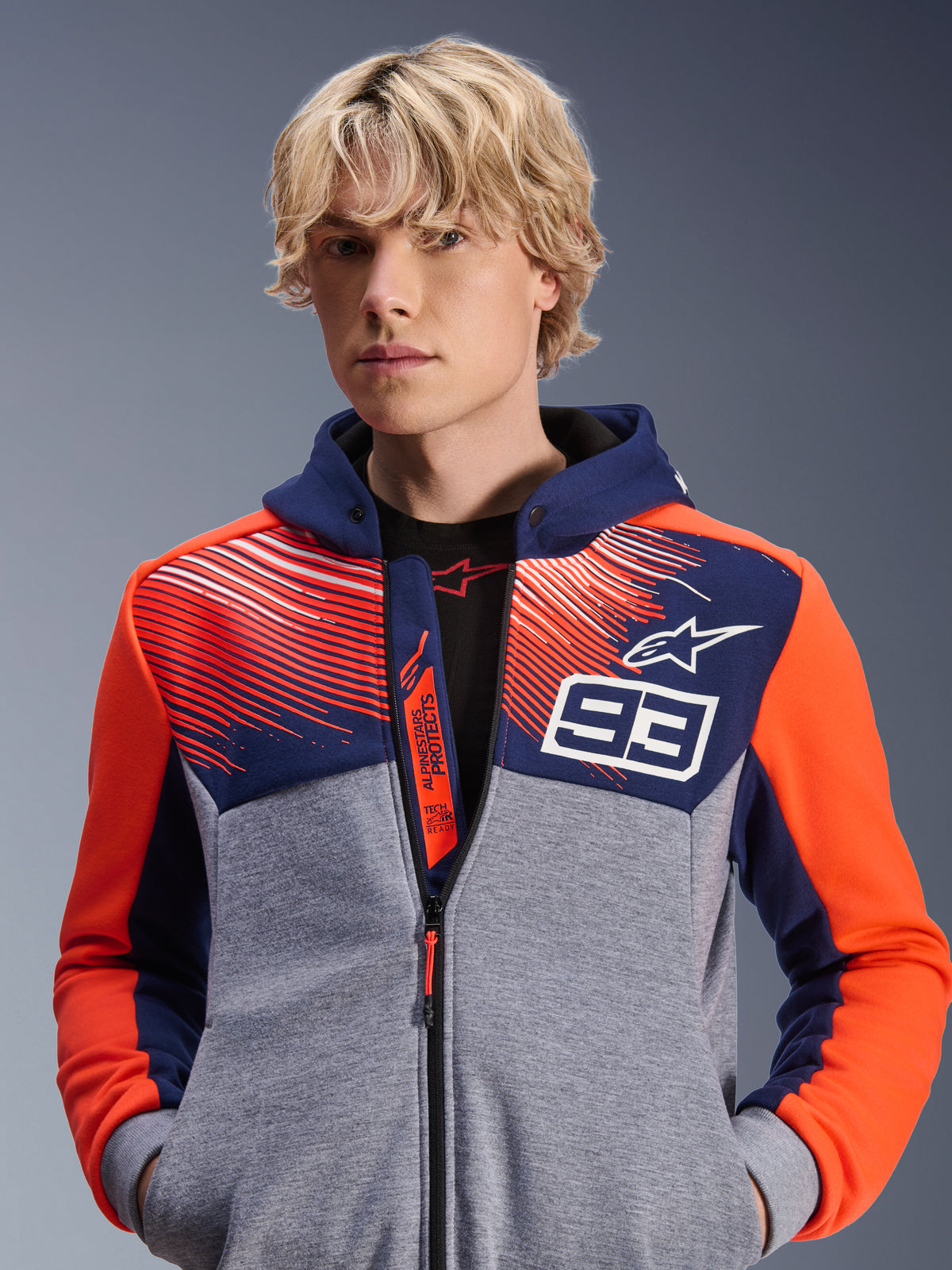 Alpinestars MM93 Chrome V2 Sport Hoodie, stedelijke motorhoodie, Blue Navy en Gray Mel met Red Fluo accenten, voorzien van Marc Marquez 93-branding en Alpinestars-logo, ontwerp met volledige ritssluiting, casual lifestyle kleding voor motorrijders, model draagt de hoodie