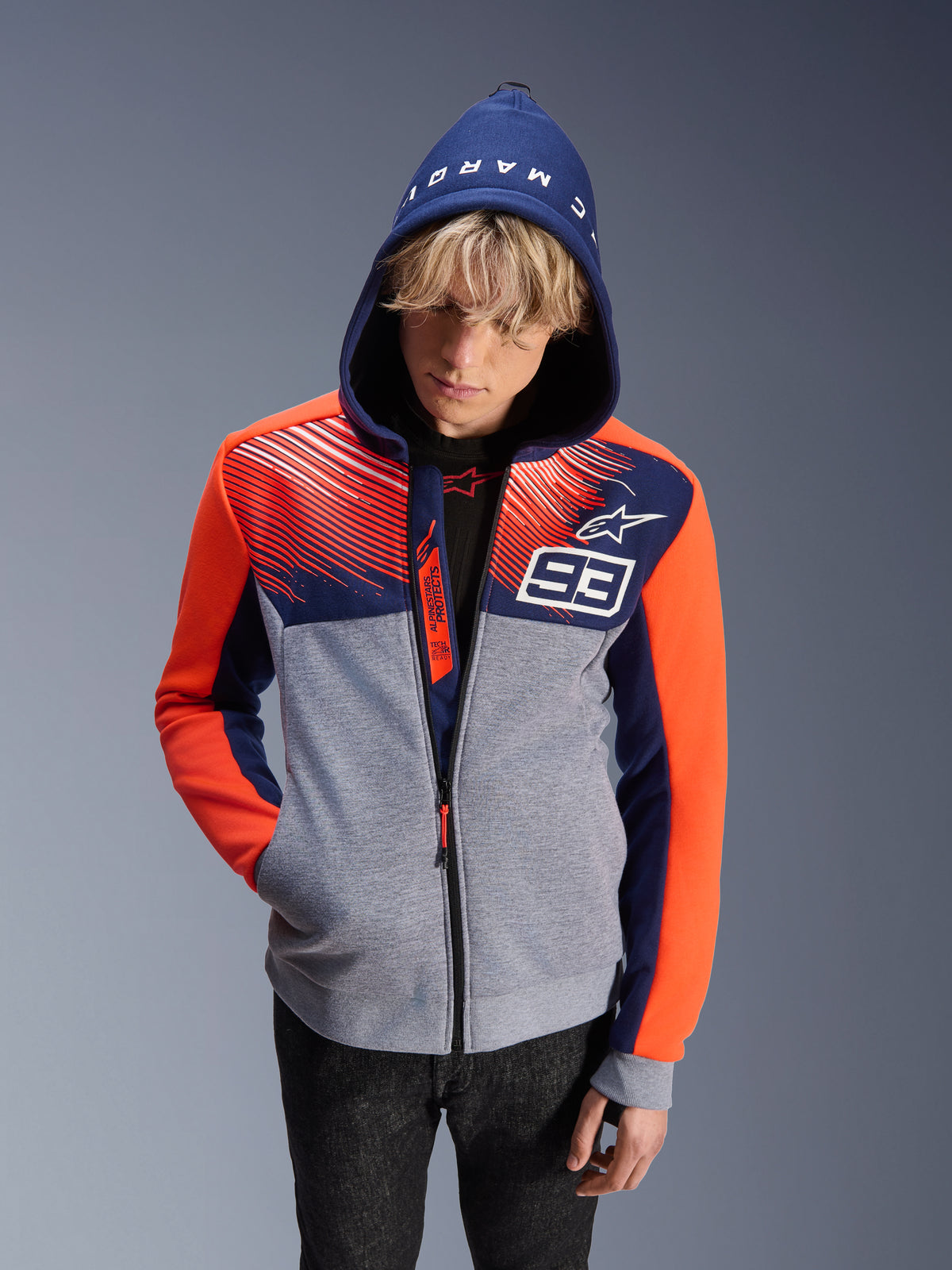 Alpinestars MM93 Chrome V2 Sport Hoodie, motorhoodie, grijs gemêleerde body met marineblauwe bovenkant en fluorescerend oranje mouwen, voorzien van het witte getal 93 en Alpinestars-sterlogo, Marc Marquez-tekst op de capuchon, gedragen door een mannelijk model met de capuchon op, urban motorsport-geïnspireerd ontwerp