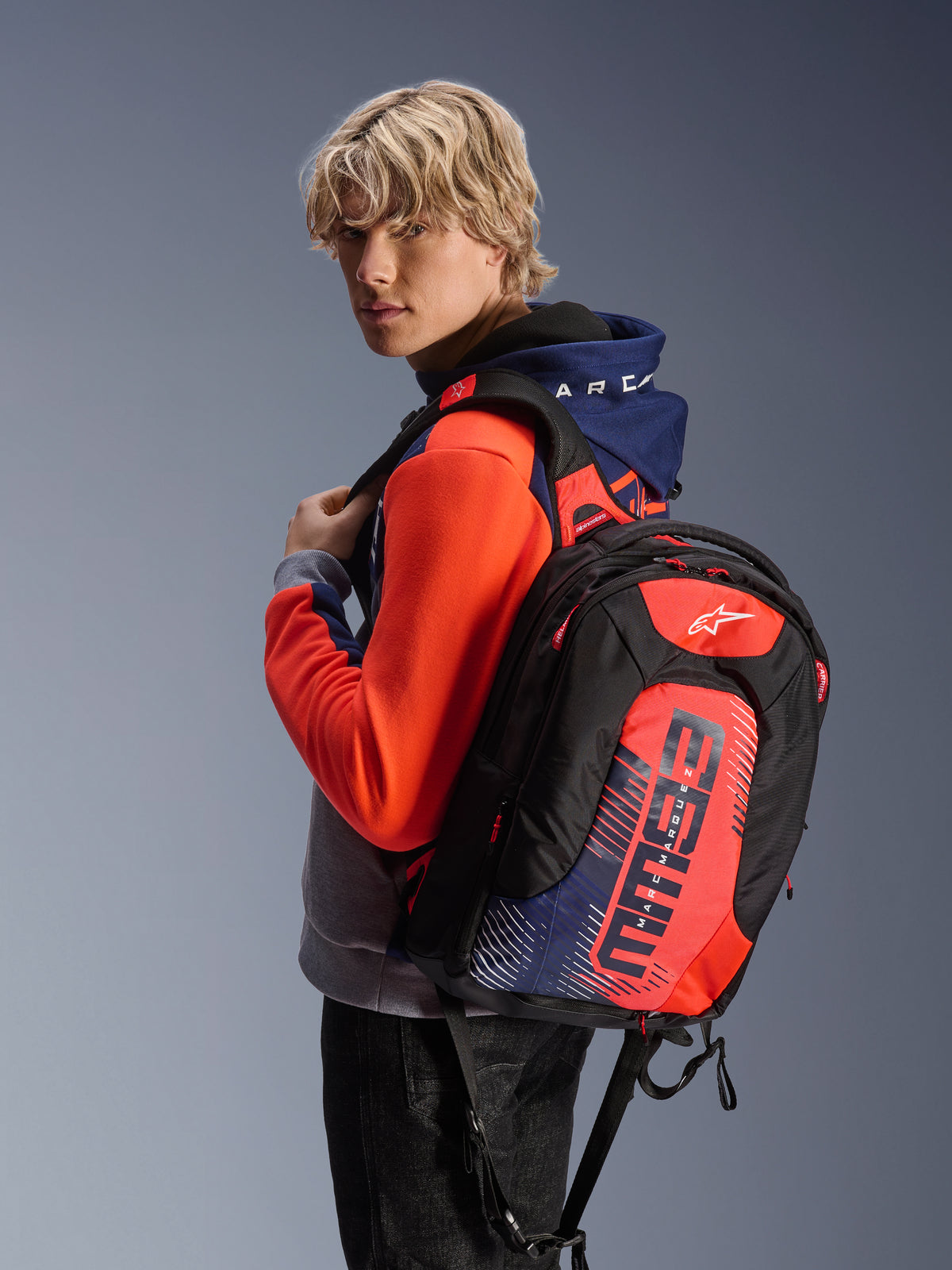 Mannelijk model draagt de Alpinestars MM93 Chrome V2 Sport Hoodie in Blue Navy Gray Mel en Red Fluo, gecombineerd met een bijpassende MM93 rugzak, casual stedelijke motorkleding met een modern atletisch silhouet.