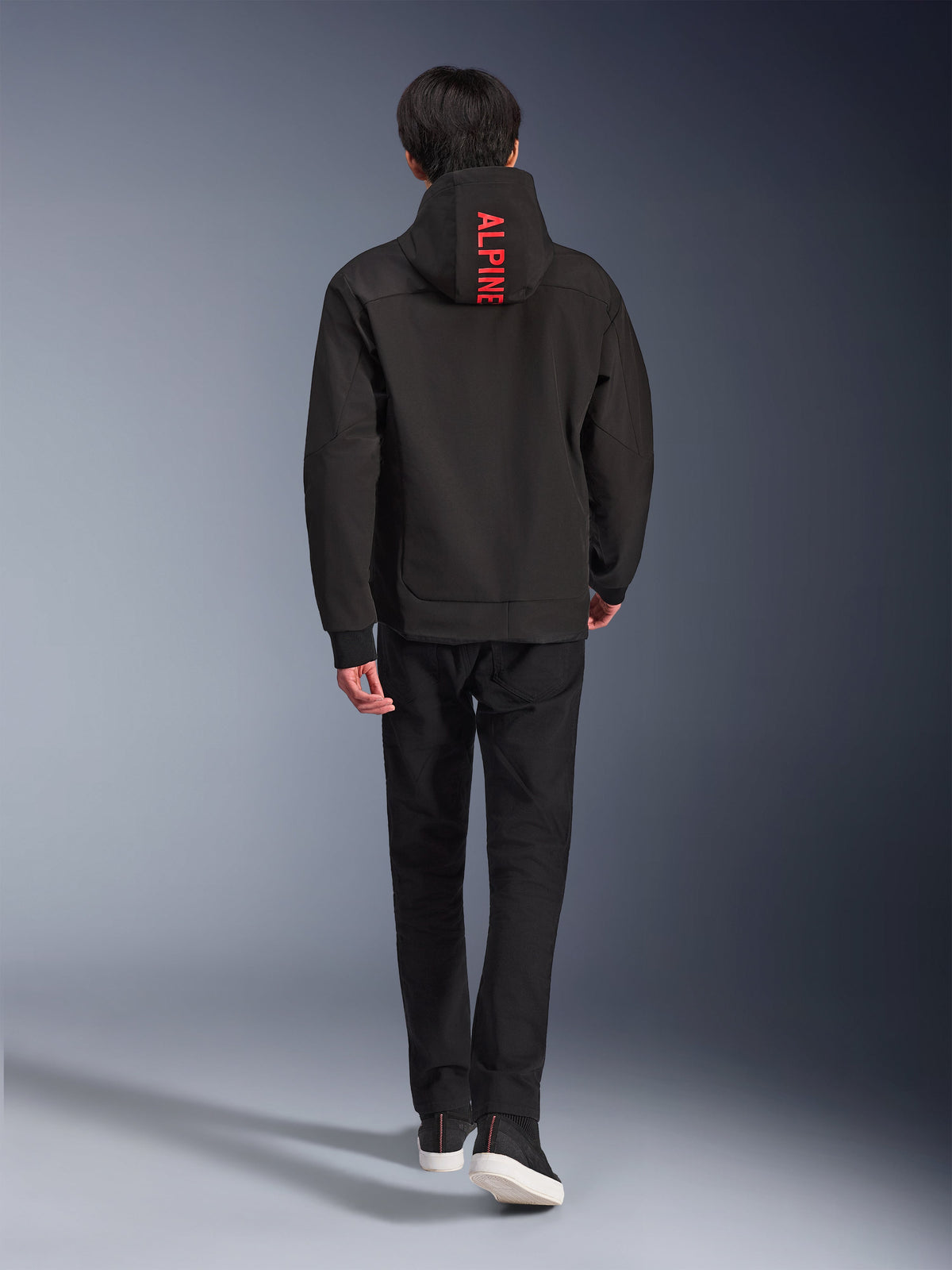 Alpinestars Radium Tech Hoodie, urban motorjas, zwart, achteraanzicht met felrode 'ALPINE'-tekst op de capuchon, gedragen met zwarte broek en sneakers, strakke technische shell voor woon-werkverkeer.