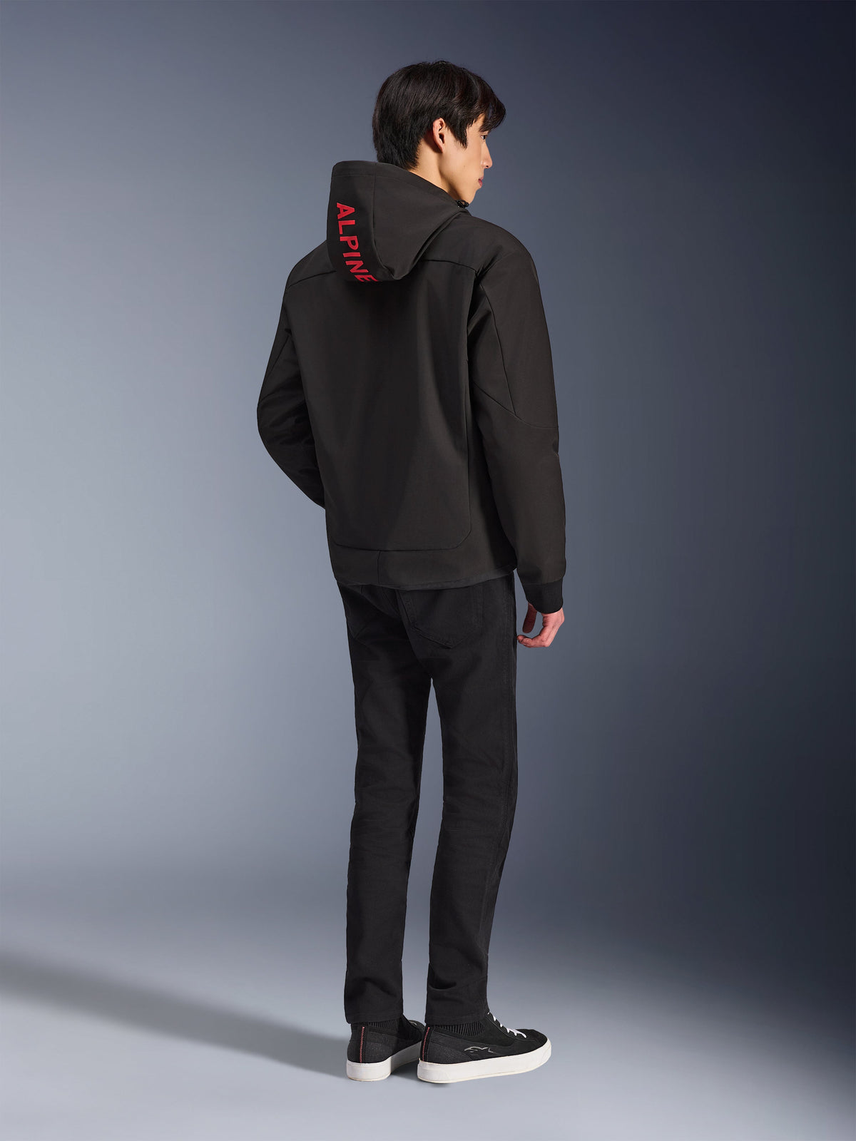 Alpinestars Radium Tech Hoodie, zwart met rode Alpinestars-branding op de capuchon, achteraanzicht op een mannelijk model, gedragen met zwarte broek en sneakers, technische stedelijke motorkleding