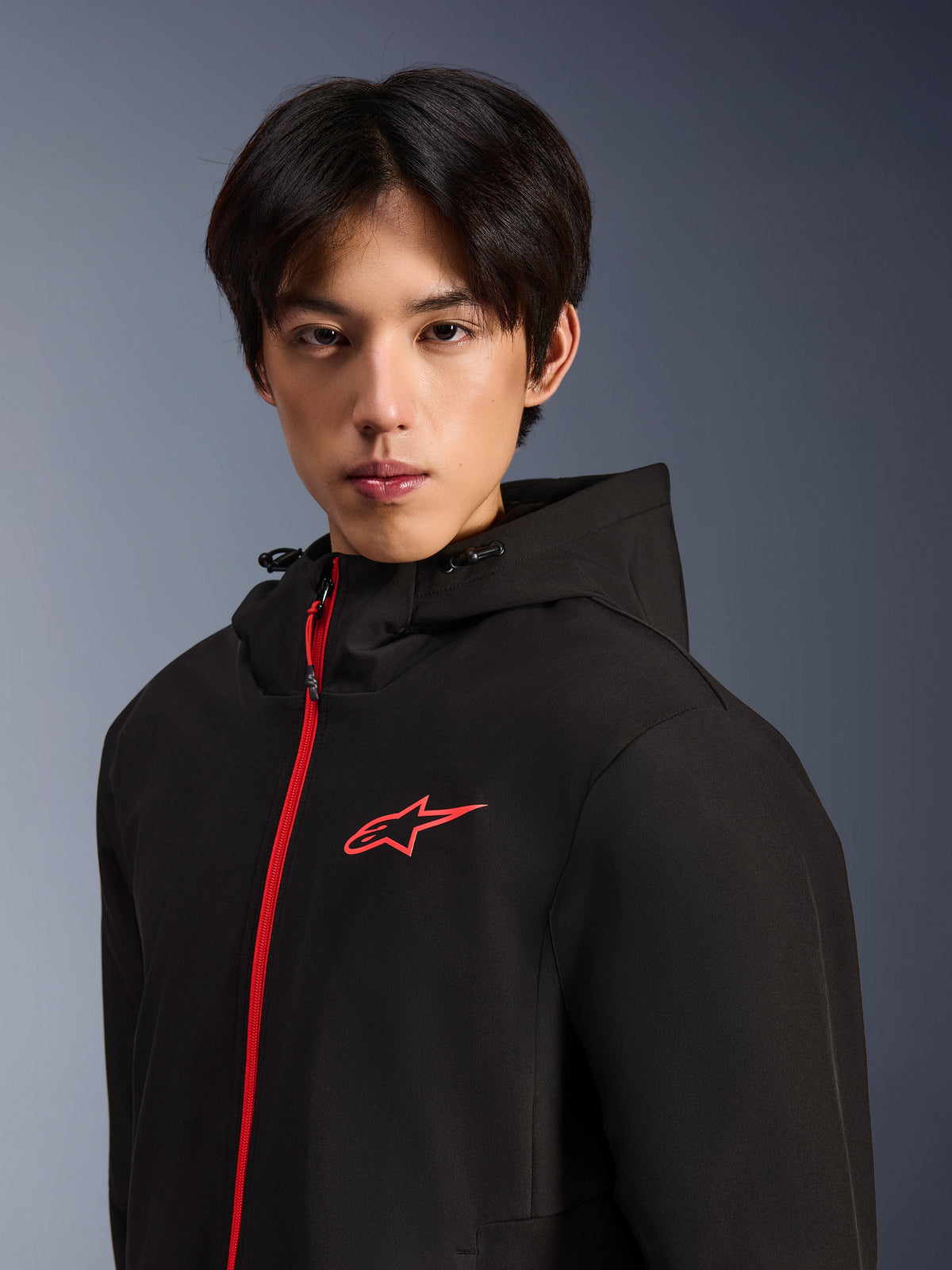Alpinestars Radium Tech Hoodie, urban motorjas, zwart en felrood, mannelijk model draagt een technische shell hoodie met een felrode rits aan de voorzijde en een logo op de borst, modern urban design
