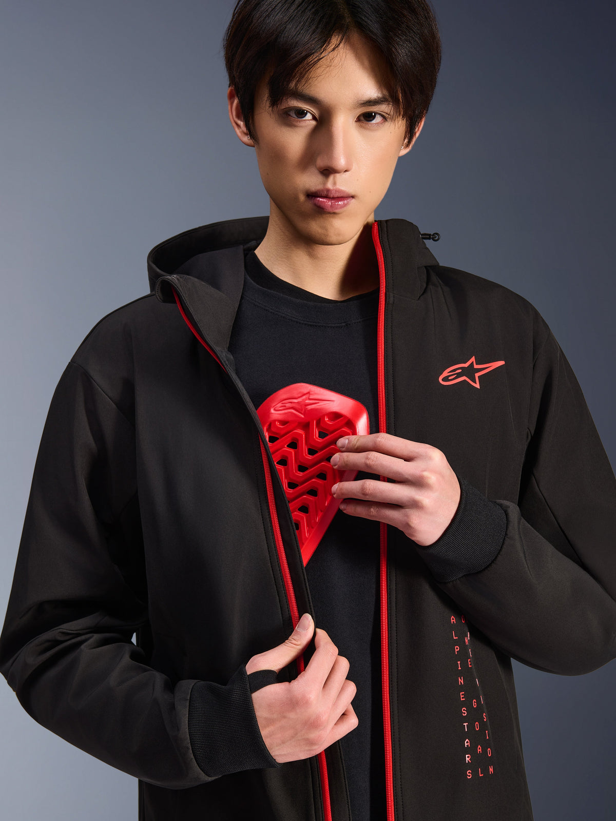 Alpinestars Radium Tech Hoodie, urban motorjas, zwart en felrood, gedragen door een model dat een rode borstbeschermer laat zien, voorzien van felrode ritsen en verticale Alpinestars-branding, strakke winddichte technische shell voor stedelijk woon-werkverkeer