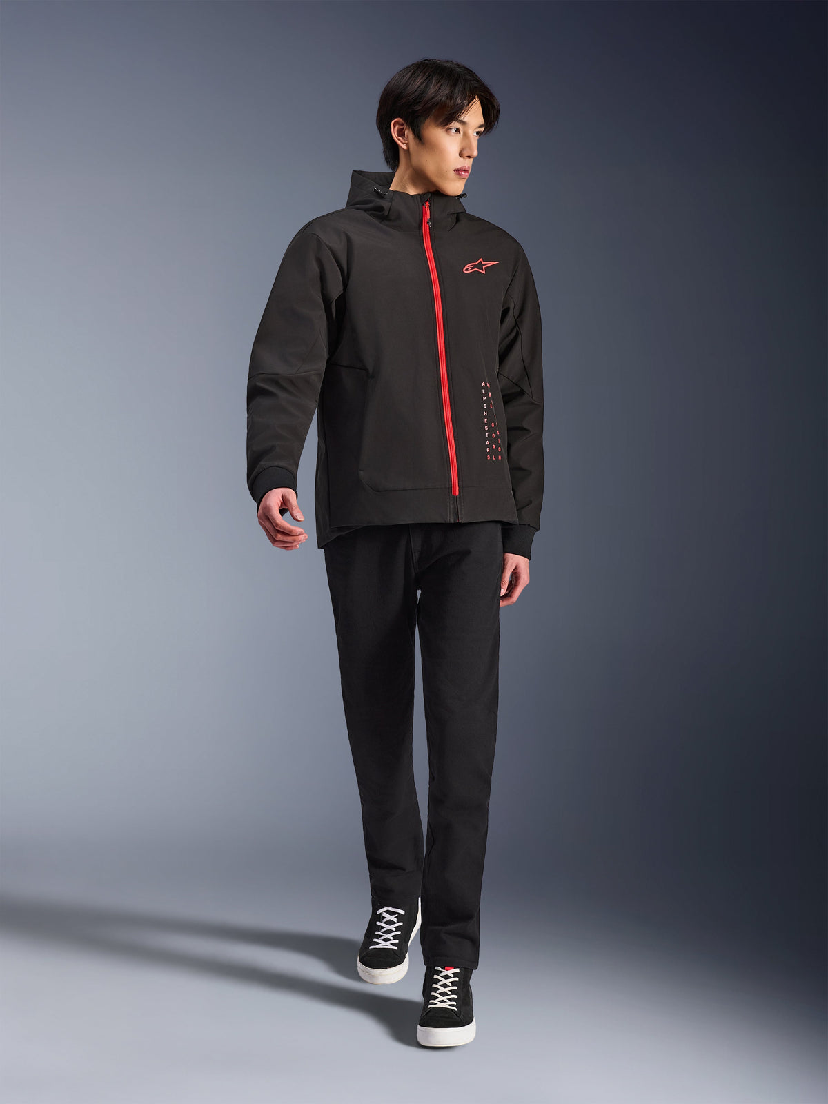 Alpinestars Radium Tech Hoodie in Black Bright Red, stedelijke motorjas gedragen door een model, voorzien van een rode ritssluiting aan de voorzijde en een logo op de borst, gecombineerd met een zwarte broek en sneakers voor een moderne woon-werk uitstraling.