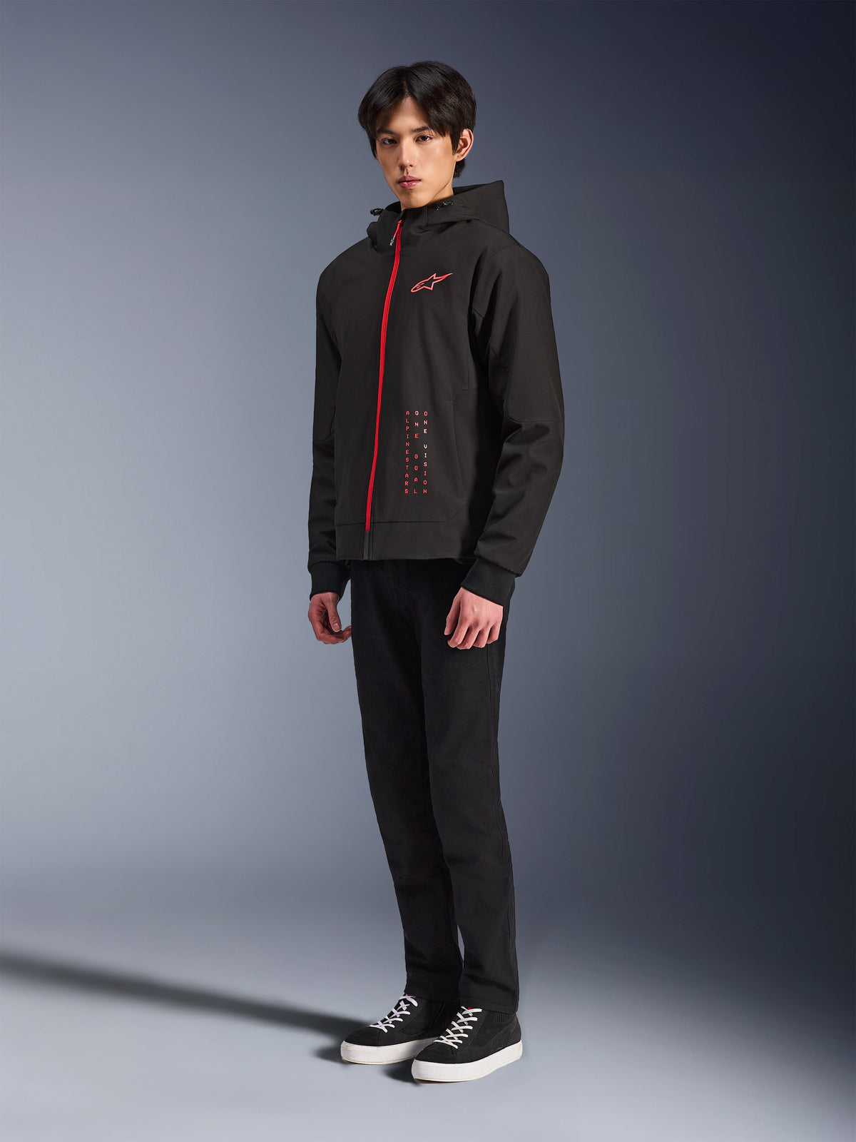 Alpinestars Radium Tech Hoodie, Black Bright Red, technische urban motorhoodie met een rode rits aan de voorzijde en een logo op de borst, gedragen door een model met zwarte broek en zwarte sneakers met witte zolen, studio-opname van het volledige lichaam