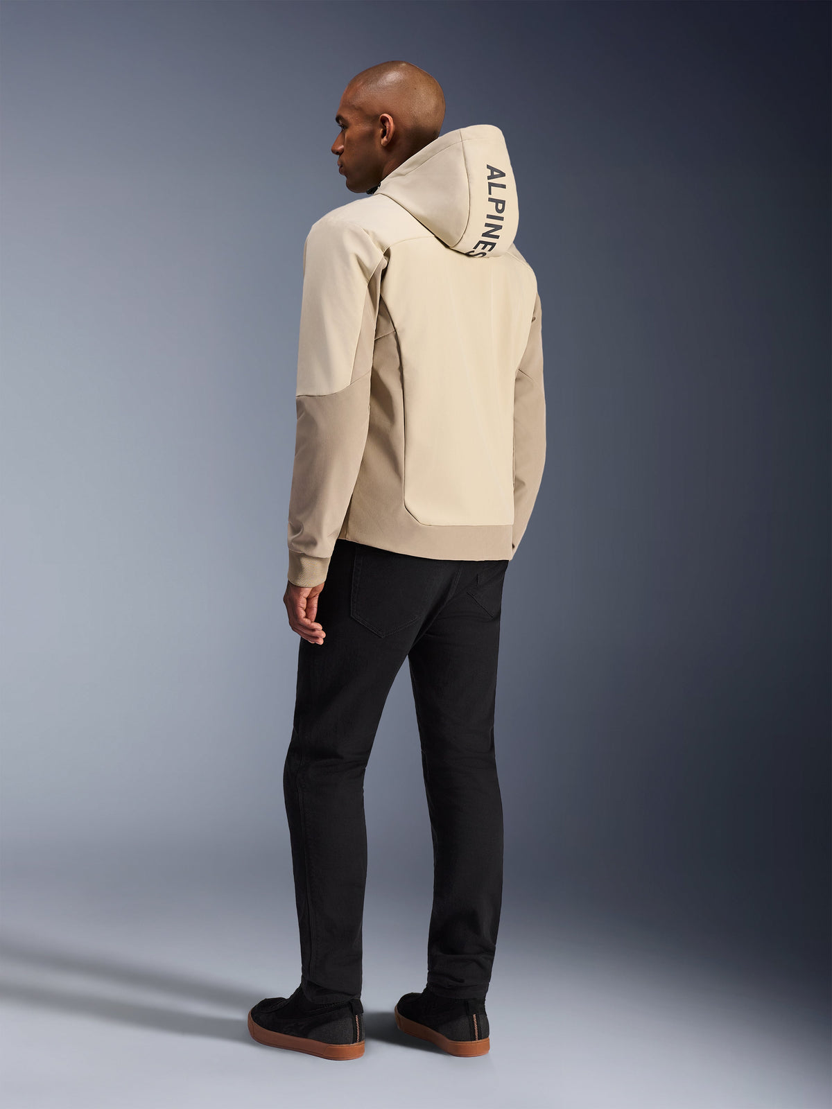 Alpinestars Radium Tech Hoodie in Mastic Aluminum, urbane Motorradjacke, Rückansicht am Model mit aufgesetzter Kapuze, vertikaler ALPINESTARS-Schriftzug auf der Kapuze, kombiniert mit schwarzer Hose, moderne Textilkonstruktion für Schutz bei Stadtfahrten