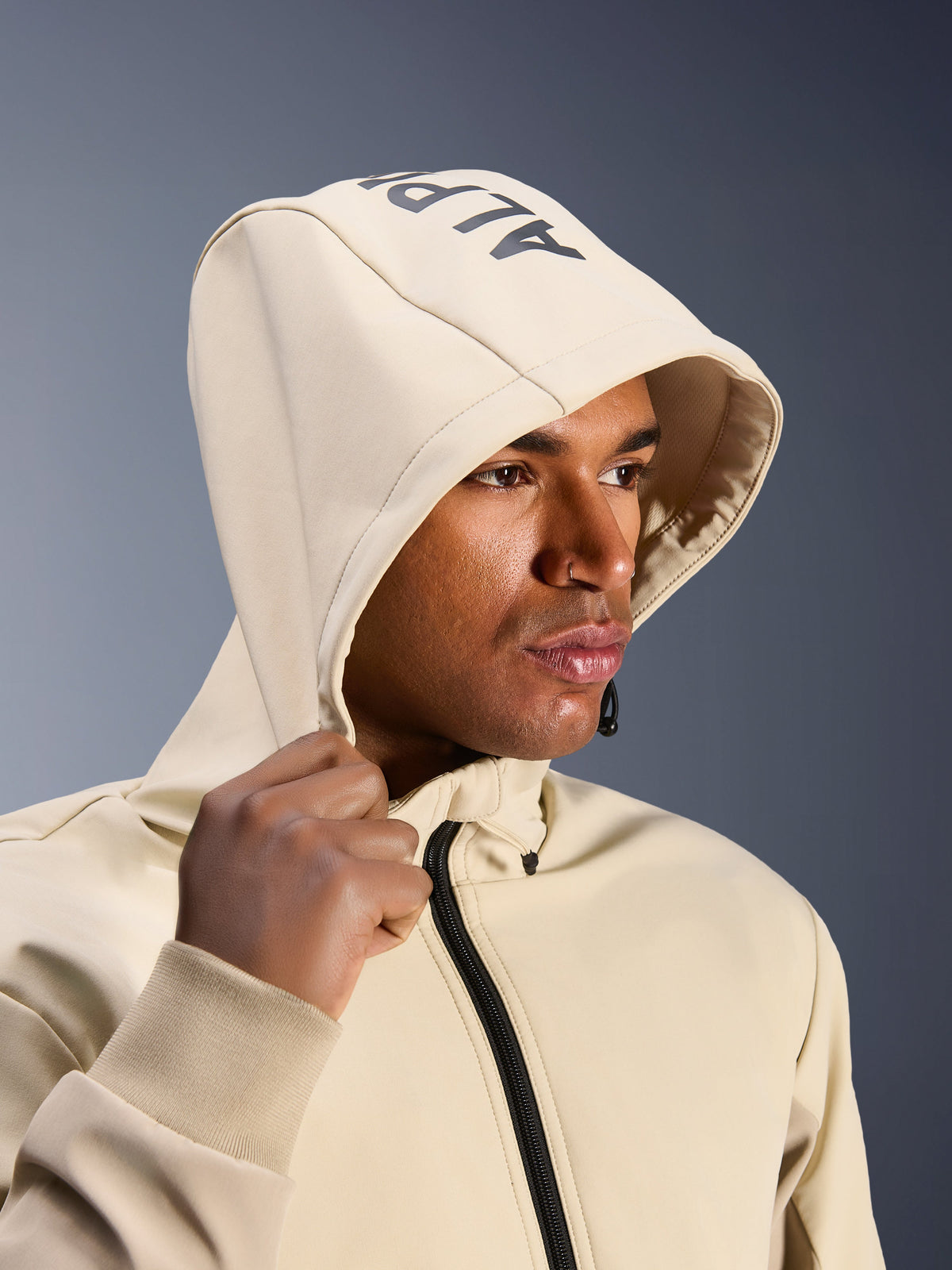 Alpinestars Radium Tech Hoodie in Mastic Aluminum, Nahaufnahme eines Mannes mit aufgesetzter Kapuze, Alpinestars-Branding auf der Oberseite, urbane Motorradjacke mit technischer winddichter Shell, moderne Streetwear-Ästhetik für Stadtfahrten