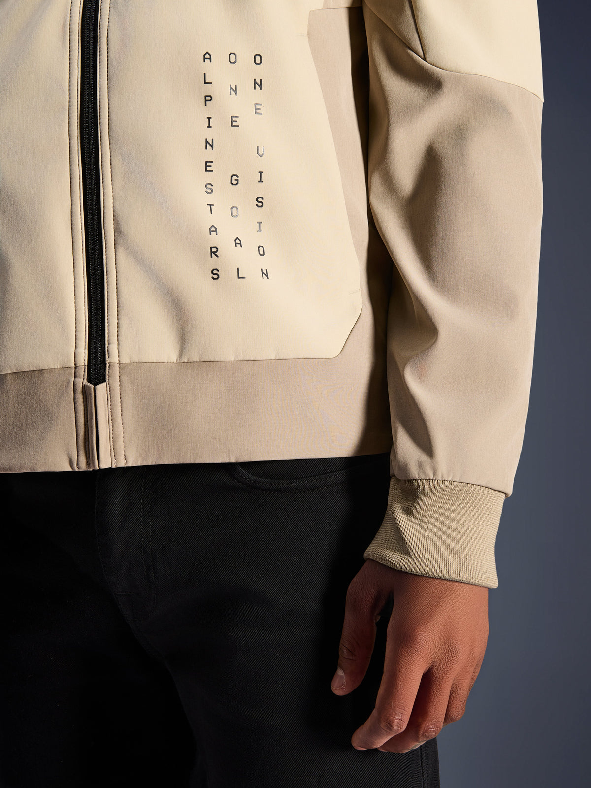 Alpinestars Radium Tech Hoodie in Mastic Aluminum, Nahaufnahme von Brust und Ärmel, vertikaler schwarzer Schriftzug 'Alpinestars One Goal One Vision', geripptes Stretch-Bündchen und Reißverschluss-Detail, urbane Motorrad-Textiljacke.