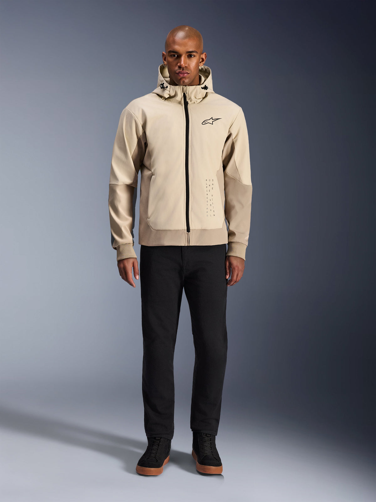 Alpinestars Radium Tech Hoodie in Mastic Aluminum, urbaner Motorrad-Hoodie, getragen von einem männlichen Model mit schwarzer Hose und schwarzen Stiefeln, mit durchgehendem schwarzem Reißverschluss, kleinem Brustlogo und athletischer Passform für Stadtfahrten.