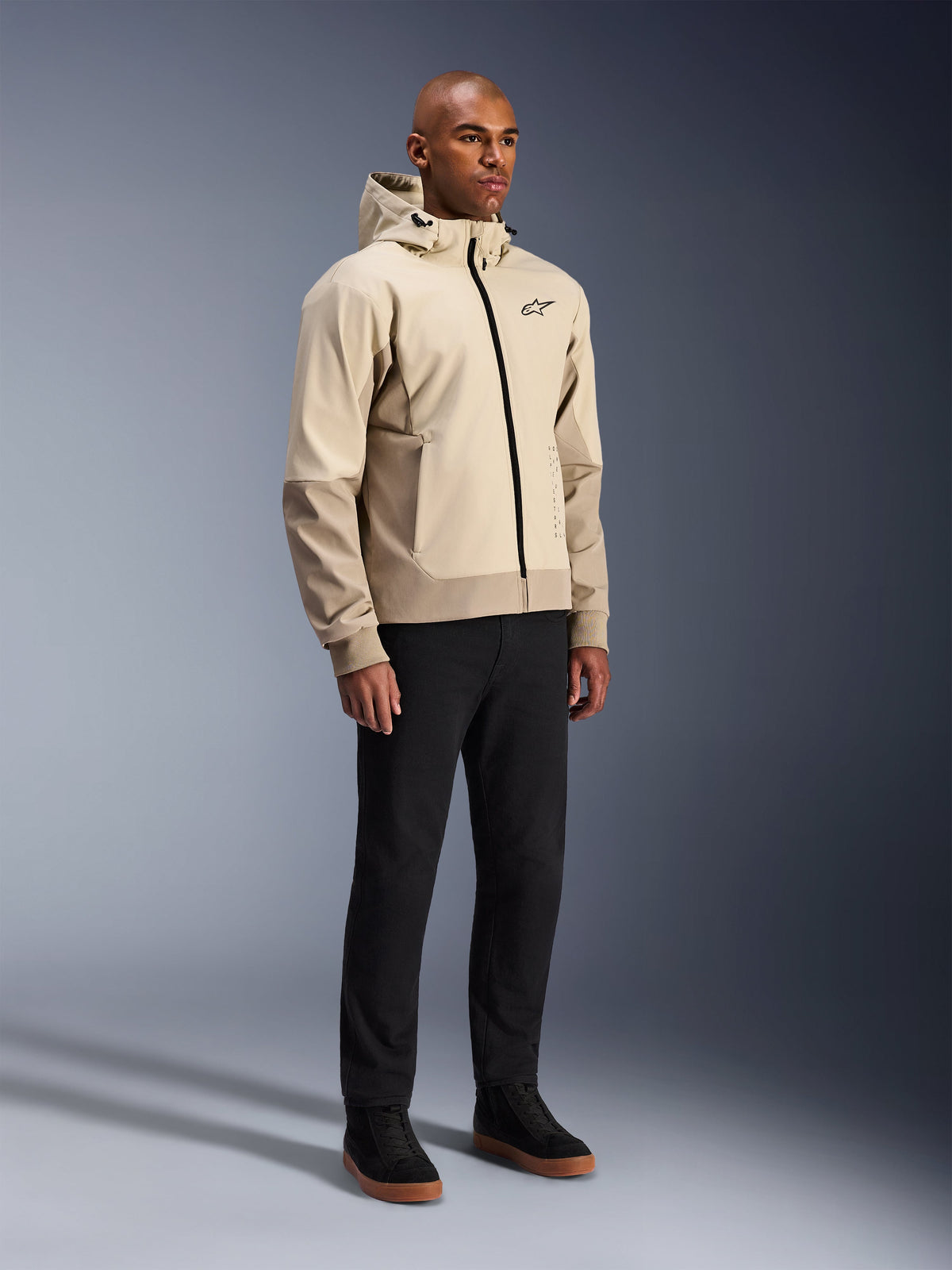 Alpinestars Radium Tech Hoodie, urbane Motorradjacke, Mastic Aluminum Hellbeige, getragen mit schwarzer Hose und schwarzen High-Top-Sneakern, mit technischer winddichter Außenschicht, Frontreißverschluss und abnehmbarer Kapuze, modernes Streetwear-Design für urbanes Fahren