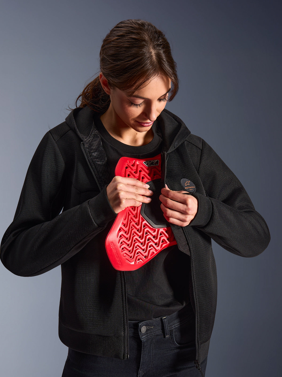 Alpinestars Stella Chrome Superairflow Hoodie, Damen-Motorrad-Hoodie, Schwarz, Modell hält einen roten Nucleon Brustprotektor, leichte 3D-Mesh-Konstruktion für urbanes und sportliches Fahren