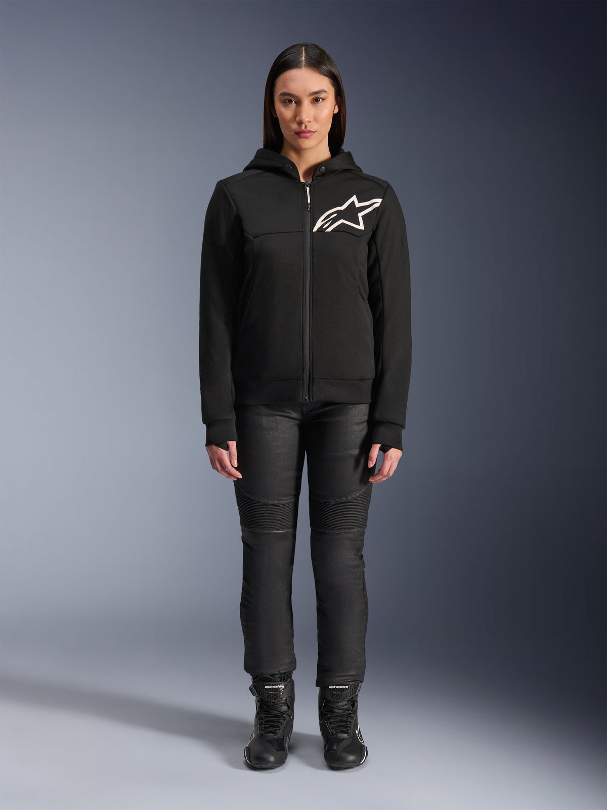 Woman wearing Alpinestars Stella Chrome V2 Sport Hoodie, Black with white Astars logo on the chest, paired with black technical riding pants and boots, urban motorcycle apparel design.