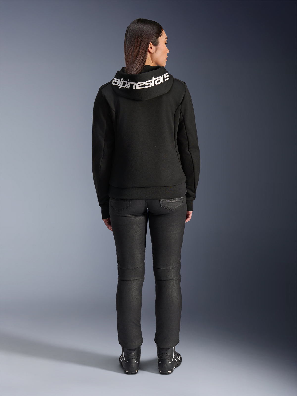 Rear view of a model wearing the Alpinestars Stella Chrome V2 Sport Hoodie in black, featuring a prominent white Alpinestars logo across the hood, paired with black riding pants and motorcycle boots, urban sport motorcycle gear for women