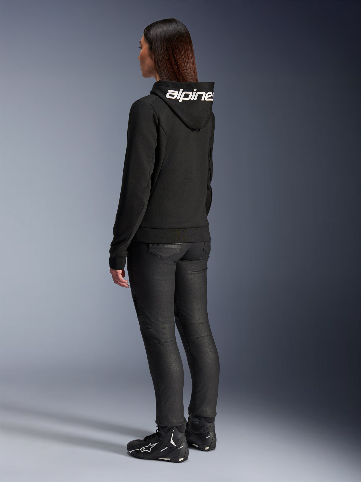Woman wearing Alpinestars Stella Chrome V2 Sport Hoodie, rear view, black motorcycle hoodie with white Alpinestars logo on the hood, paired with black technical riding pants and black motorcycle shoes, casual urban commuting gear