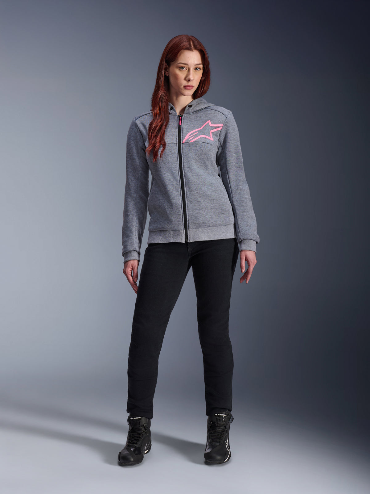 Alpinestars Stella Chrome V2 Sport Hoodie, gris chiné rose fluo, porté par une femme avec un pantalon et des bottes noirs, arborant un logo étoile Alpinestars rose sur la poitrine, équipement de moto urbain entièrement zippé, polaire technique légère avec une coupe athlétique décontractée.