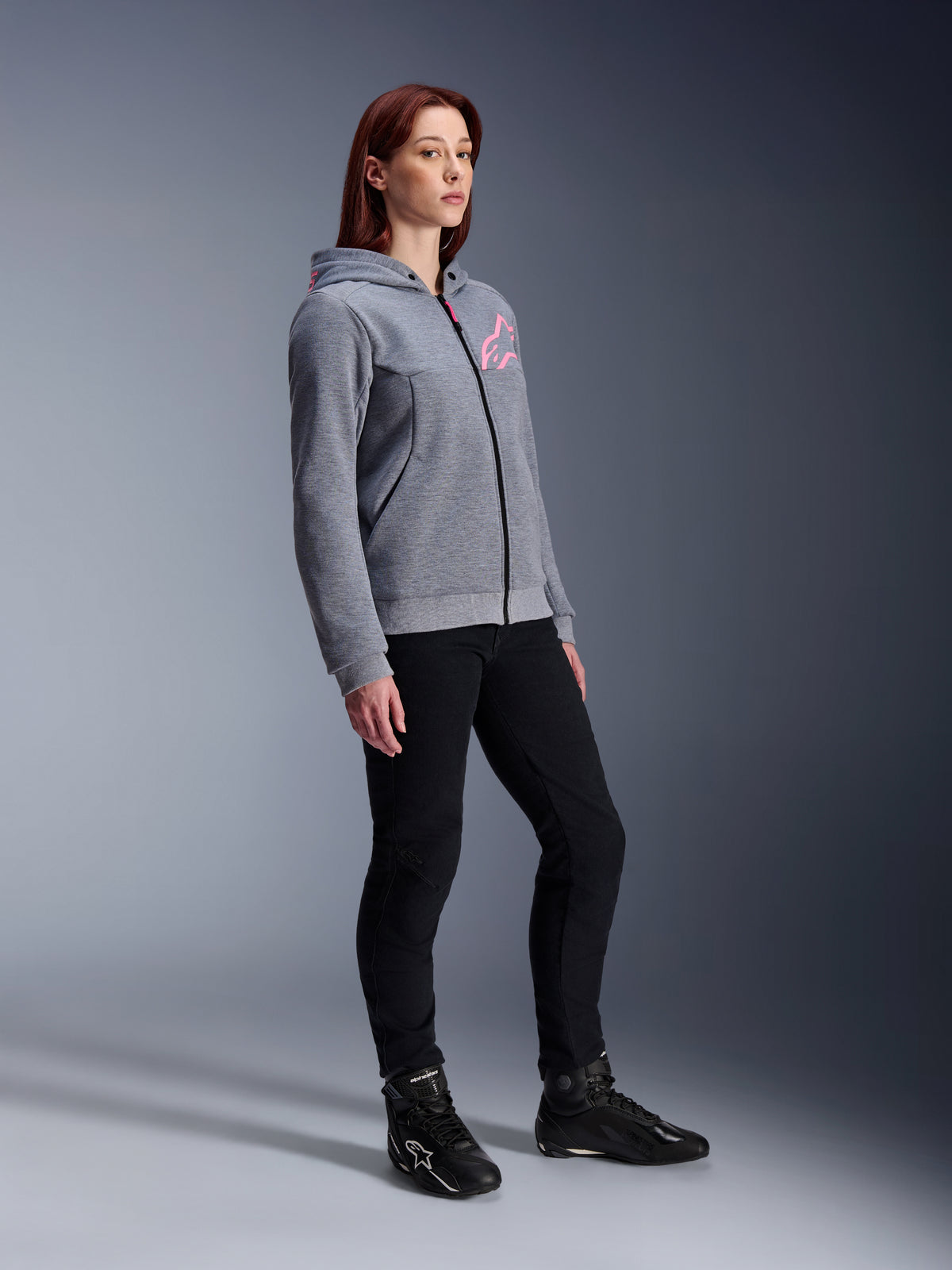 Alpinestars Stella Chrome V2 Sport Hoodie, sweat à capuche de moto pour femme, gris chiné et rose fluo, porté avec un pantalon de moto noir et des bottines noires, équipement de conduite urbain décontracté