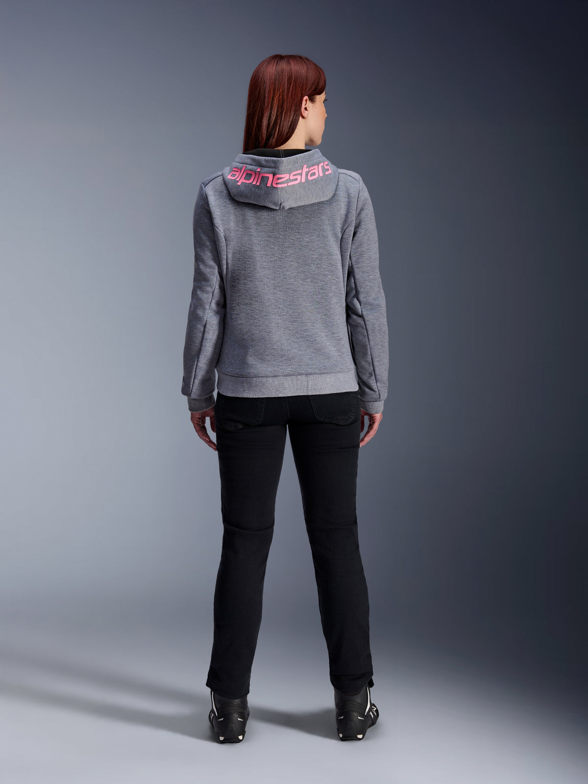 Sweat à capuche sport Stella Chrome V2 Alpinestars, sweat à capuche de moto pour femme, gris chiné et rose fluo, vue arrière montrant le logo Alpinestars rose sur la capuche, porté avec un pantalon de moto noir et des bottes, vêtement de moto décontracté pour femme