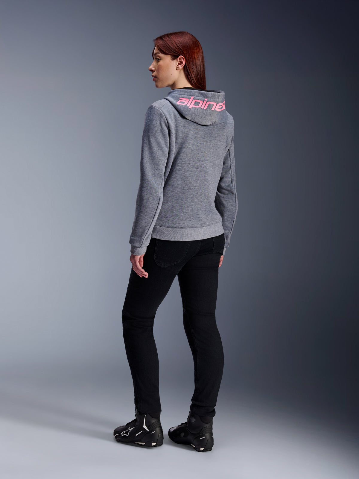 Sweat à capuche sport Alpinestars Stella Chrome V2, sweat à capuche moto pour femme, gris chiné avec marquage rose fluo sur la capuche, vue arrière, porté avec un pantalon noir et des chaussures de moto, style urbain décontracté avec protection moto