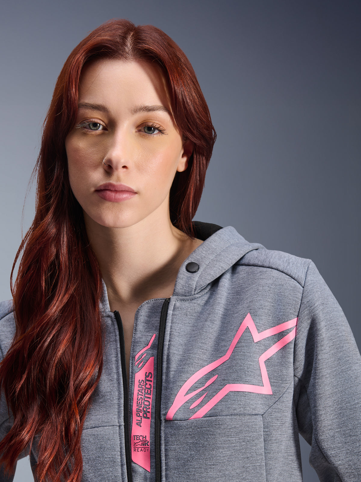 Femme portant le sweat à capuche Alpinestars Stella Chrome V2 Sport, Gris Chiné Rose Fluo, gros plan montrant le logo étoile Alpinestars rose et le marquage Tech-Air Ready, vêtement de moto urbain.