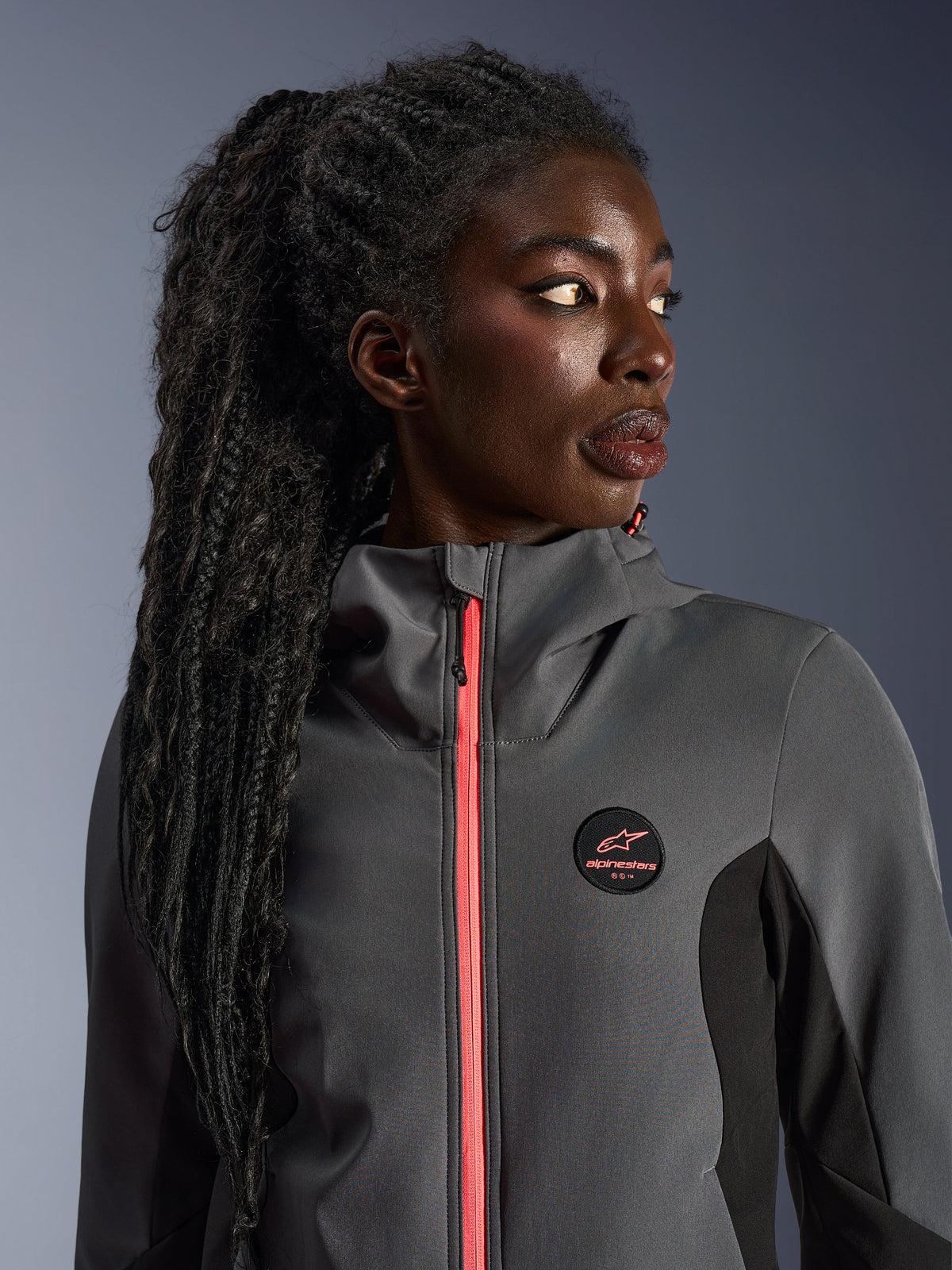 Female model wearing the Alpinestars Stella Radium Tech Hoodie in deep gray and black, featuring a bright coral-colored zipper, circular chest logo, and high-neck windproof shell, casual urban motorcycle gear for women
