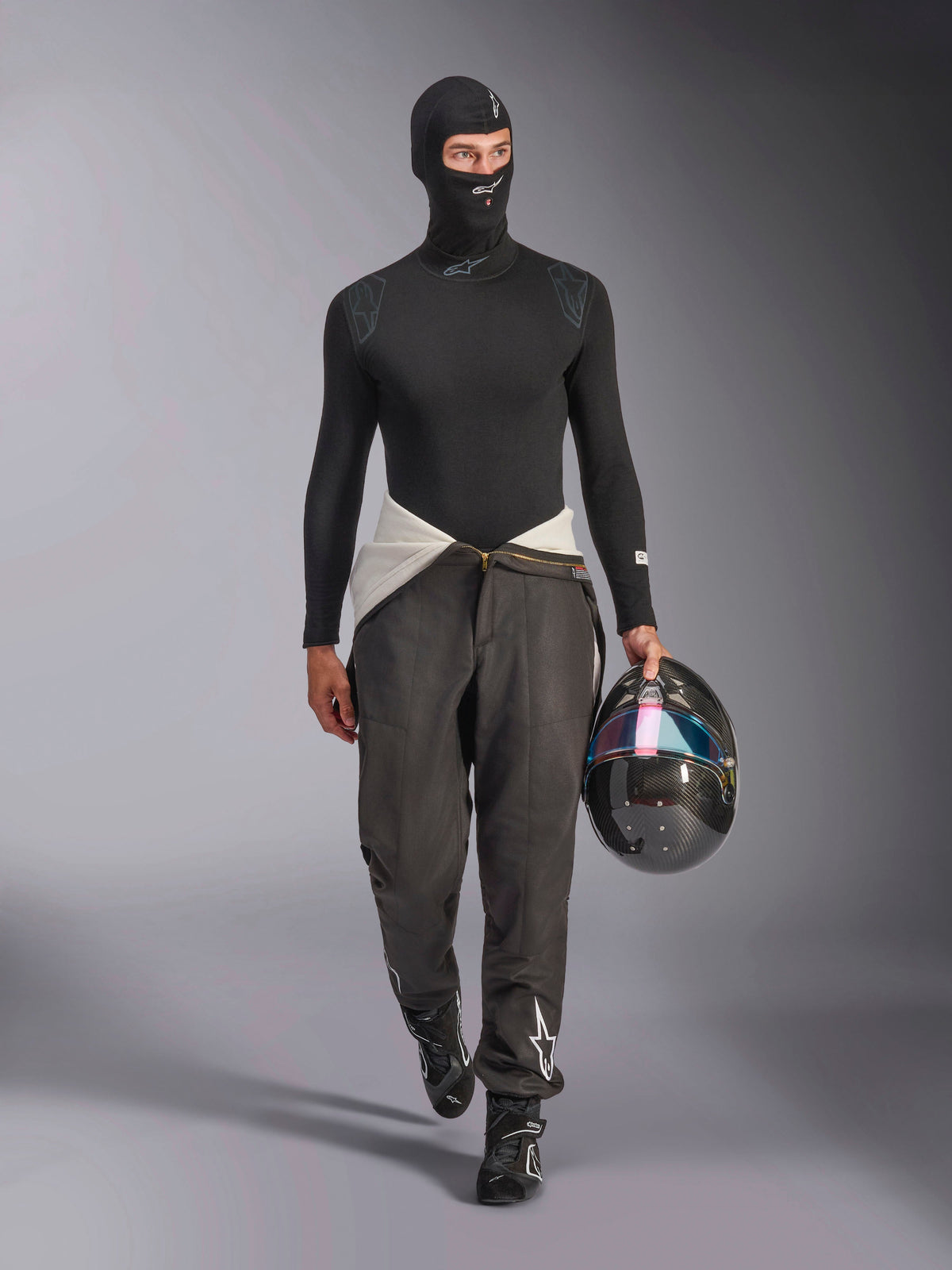 Alpinestars ZX Evo V3 Bivakmuts, zwart, gedragen door een model in een volledige race-onderkleding outfit inclusief een bijpassend shirt met lange mouwen en een grijze racebroek, houdt een carbon helm vast met een iriserend vizier, professionele autorace-uitrusting