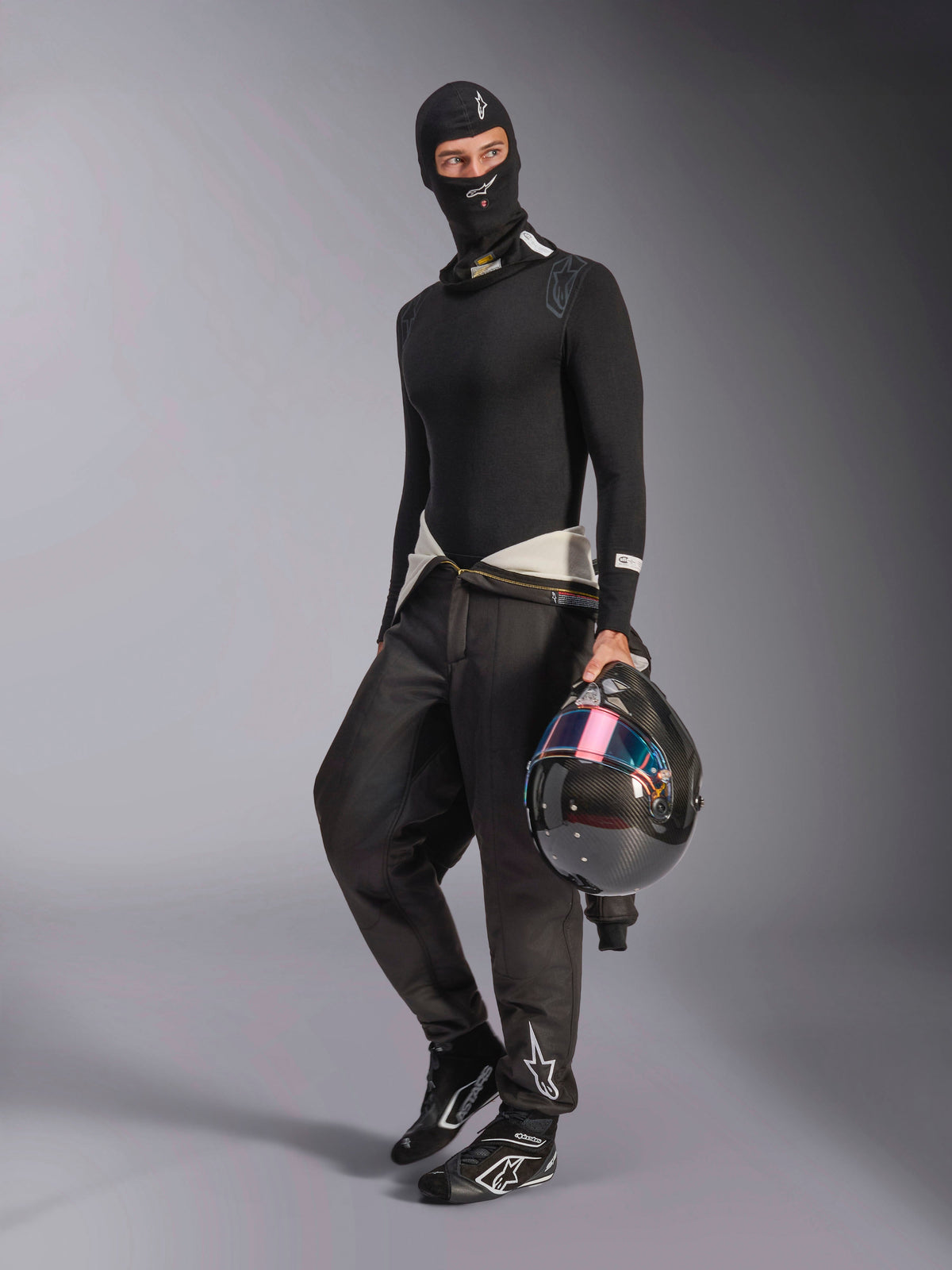 Alpinestars ZX Evo V3 Bivakmuts, zwart, gedragen door een coureur met volledige auto racing basislaag uitrusting, voorzien van het witte logo op het voorhoofd, vasthouden van een koolstofvezel helm met een iriserend vizier, professionele brandwerende motorsportkleding
