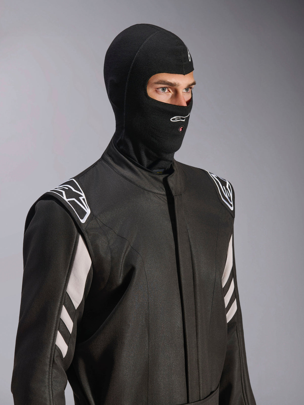 Alpinestars ZX Evo V3 Bivakmuts, zwart, vuurbestendige hoofdbescherming voor autoracen, gedragen door een model in een zwart racepak met witte accenten, voorzien van Alpinestars-logo op de kin, opening voor hydratatieslang, professionele beschermende uitrusting voor de autosport