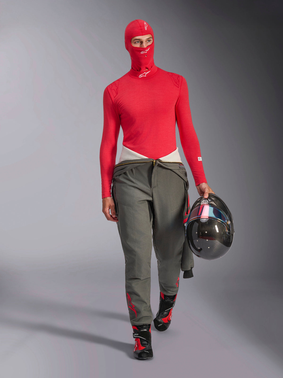 Pilote de course portant la cagoule Alpinestars ZX Evo V3 rouge, sous-vêtement technique à manches longues rouge assorti, pantalon de course gris, bottes de course noires et rouges, tenant un casque en fibre de carbone, ensemble d'équipement de course automobile professionnel