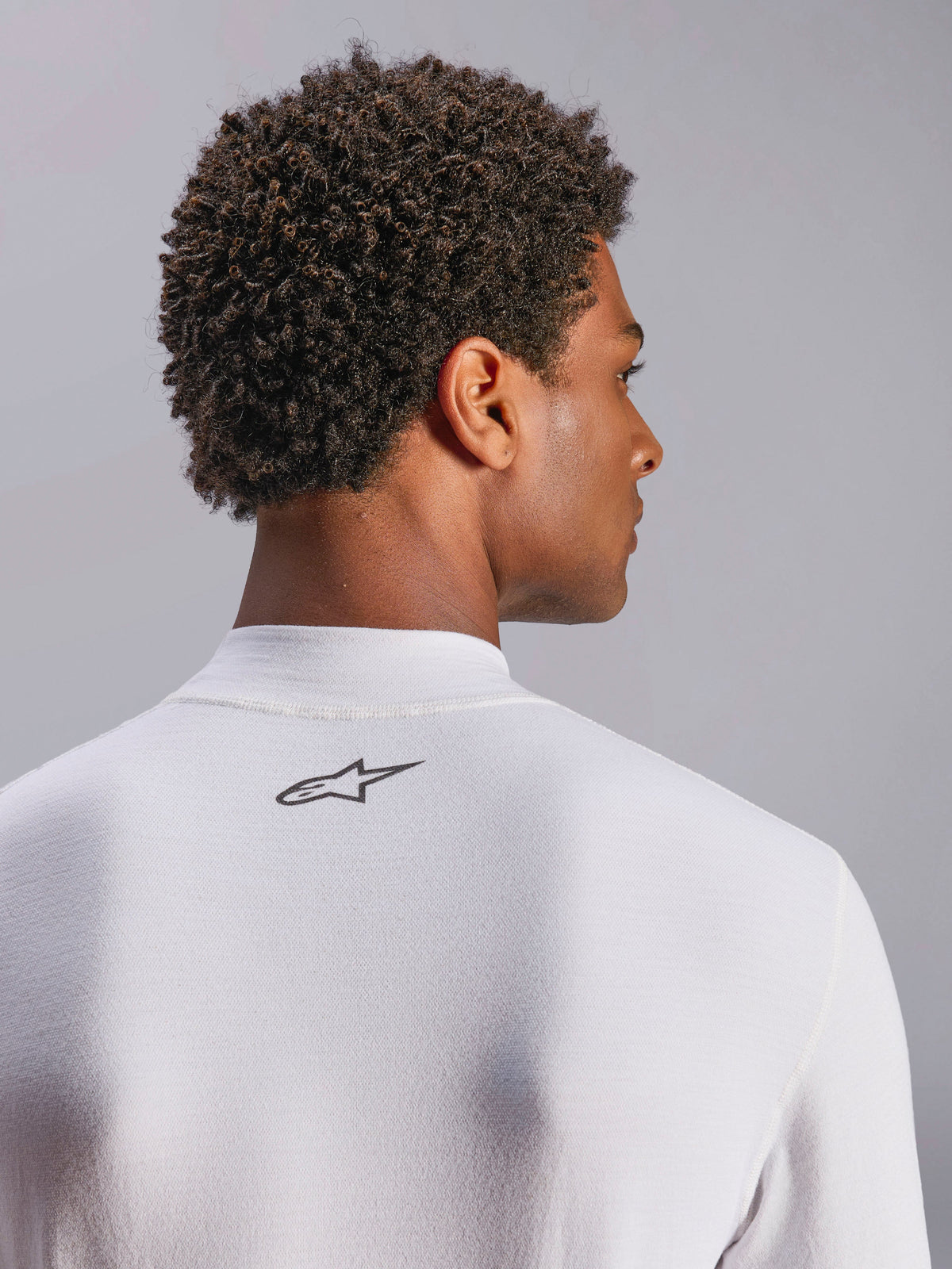Haut Alpinestars ZX Evo V3, sous-vêtement de course automobile à manches longues, blanc, gros plan sur la zone de la nuque avec logo Alpinestars noir, tissu technique résistant aux flammes, porté par un mannequin aux cheveux sombres et bouclés