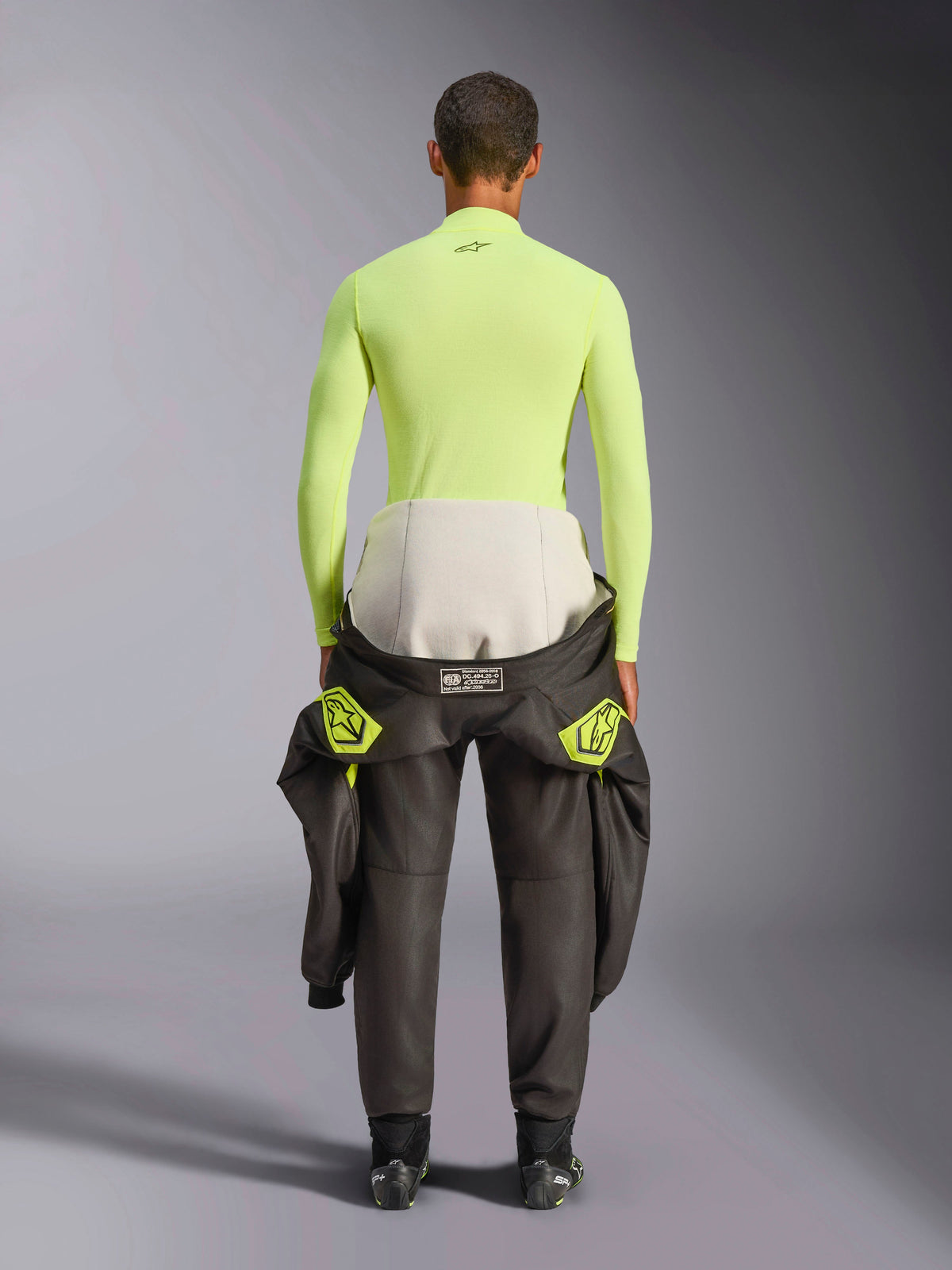 Haut Alpinestars ZX Evo V3, sous-vêtement technique à manches longues, jaune fluo, vue arrière d'un pilote avec une combinaison de course nouée autour de la taille, haut résistant aux flammes jaune néon éclatant avec un petit logo noir sur le col et une coupe athlétique ajustée