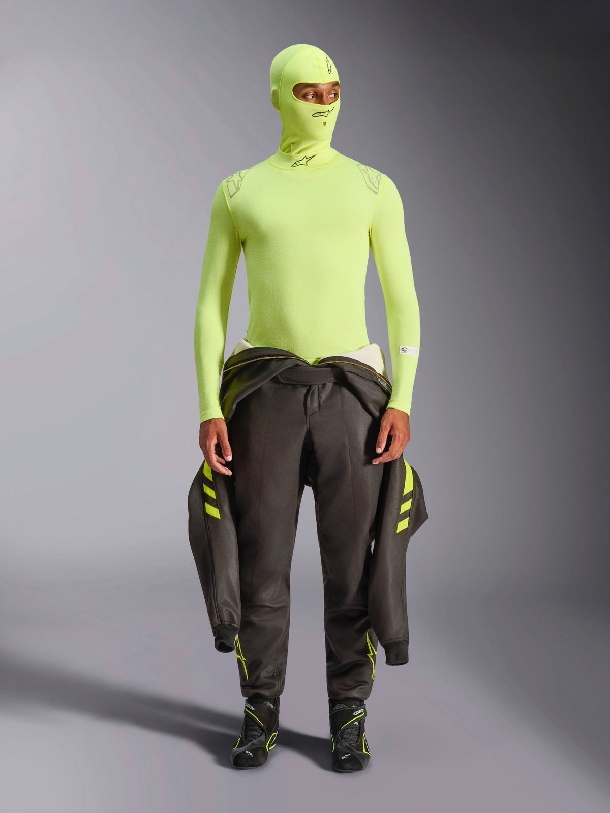 Haut à manches longues Alpinestars ZX Evo V3, Jaune Fluo, porté avec une cagoule assortie et une combinaison de course sombre nouée à la taille, sous-vêtement technique ignifugé léger pour la course automobile, logos Alpinestars sur le col et les épaules