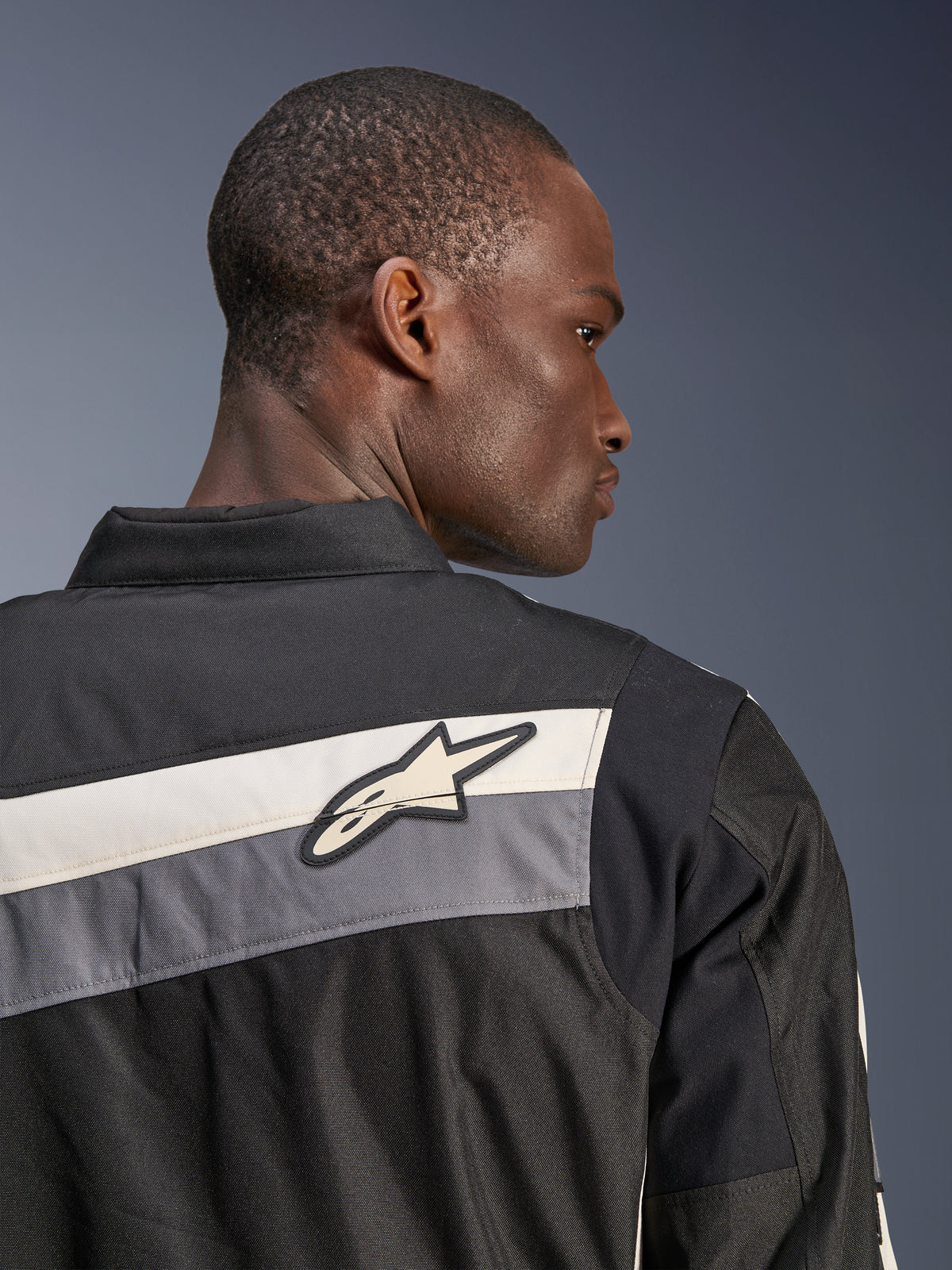 Alpinestars T-Dyno WR Jas, textiele motorjas, zwart, donkergrijs en ecru, close-up van de bovenrug en schouder met horizontale strepen en het Alpinestars sterlogo, gedragen door een model, erfgoed-geïnspireerd ontwerp voor rijden op de weg
