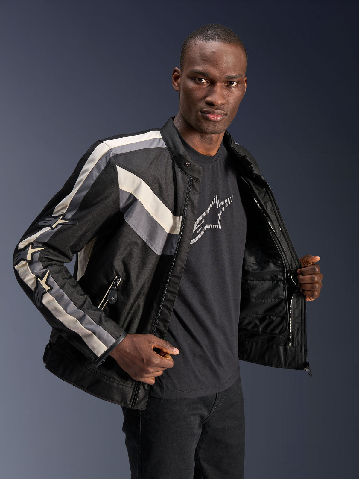 Alpinestars T-Dyno WR jas, zwart, donkergrijs en ecru, textiele motorjas met vintage-geïnspireerde biker-pasvorm, streepdetails op de borst en mouwen, gedragen over een zwart grafisch T-shirt, urban motoruitrusting