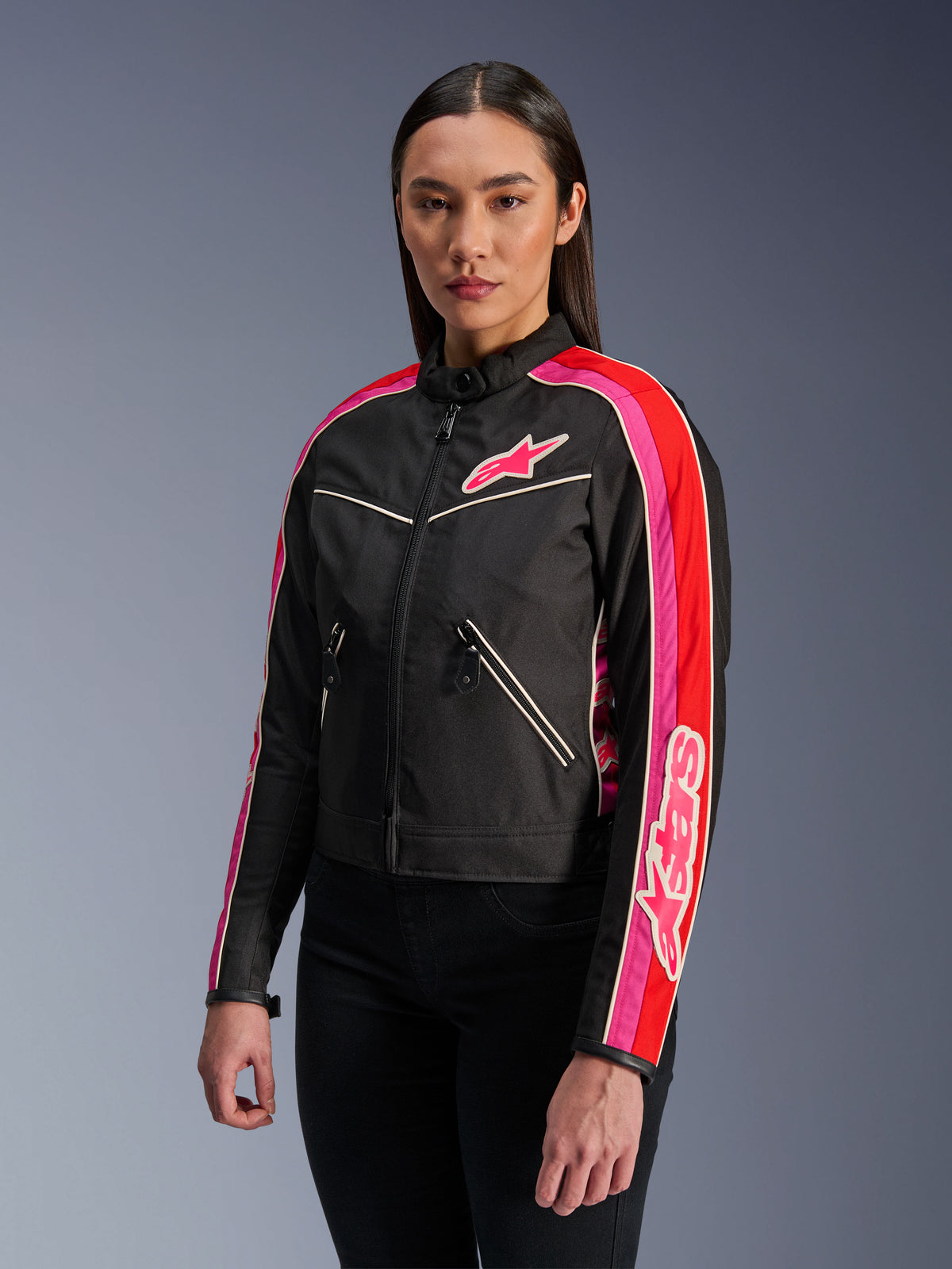 Vrouw draagt de Alpinestars Stella T-Dyno WR Jas in Black Bright Red Fuchsia, textiele motorjas met een vintage-geïnspireerde biker-pasvorm, voorzien van opvallende fuchsia en rode strepen op de mouwen en een logo op de borst, gecombineerd met een zwarte broek.