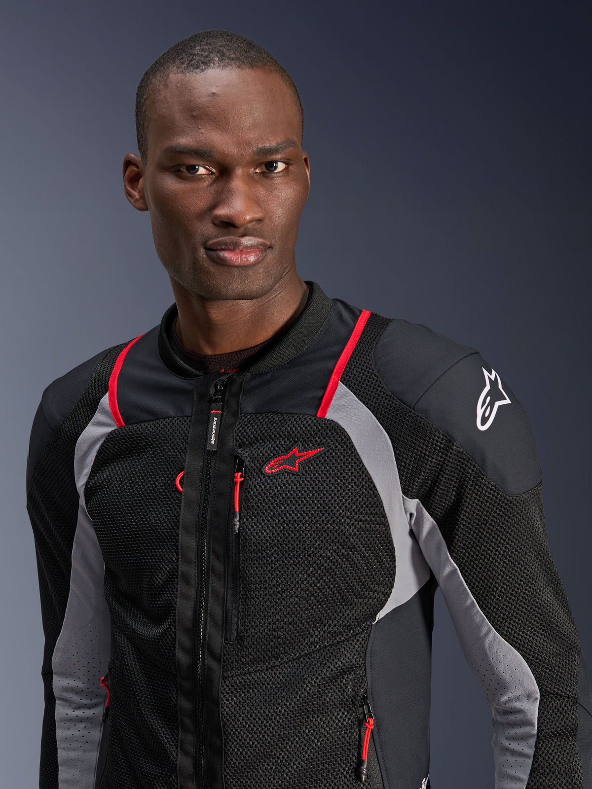 Homme portant la veste Alpinestars Troop-Air Pro, noir, gris cendre et rouge vif, veste technique ventilée avec panneaux en maille respirante, liserés rouges et logo Alpinestars sur l'épaule, équipement moto urbain contemporain