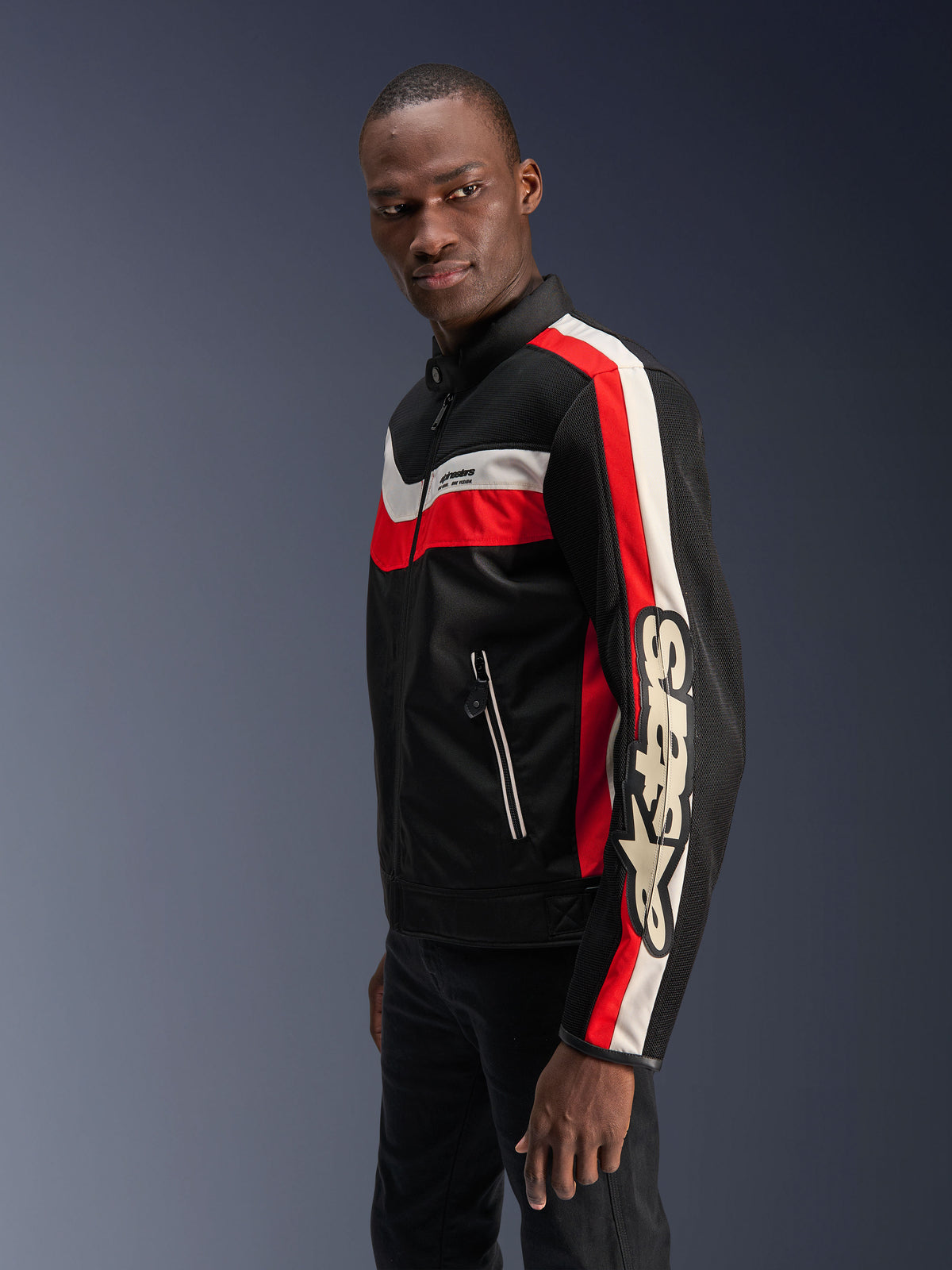 Männliches Model in der Alpinestars T-Dyno Air Jacke in Schwarz, Bright Red und Ecru, Vintage-inspirierte Textil-Motorradjacke mit 3D-Mesh-Einsätzen, markanten roten und cremeweißen Streifen sowie auffälligem Alpinestars-Branding auf dem Ärmel.