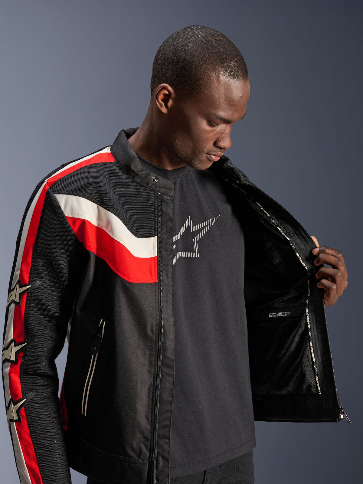 Alpinestars T-Dyno Air Jacke, Schwarz, Leuchtend Rot und Ecru, männliches Model öffnet die Jacke, um das Innenfutter und das wasserdichte Fach zu zeigen, vintage-inspirierte Textiljacke mit 3D-Mesh-Einsätzen und Stern-Logos, moderne Motorradbekleidung