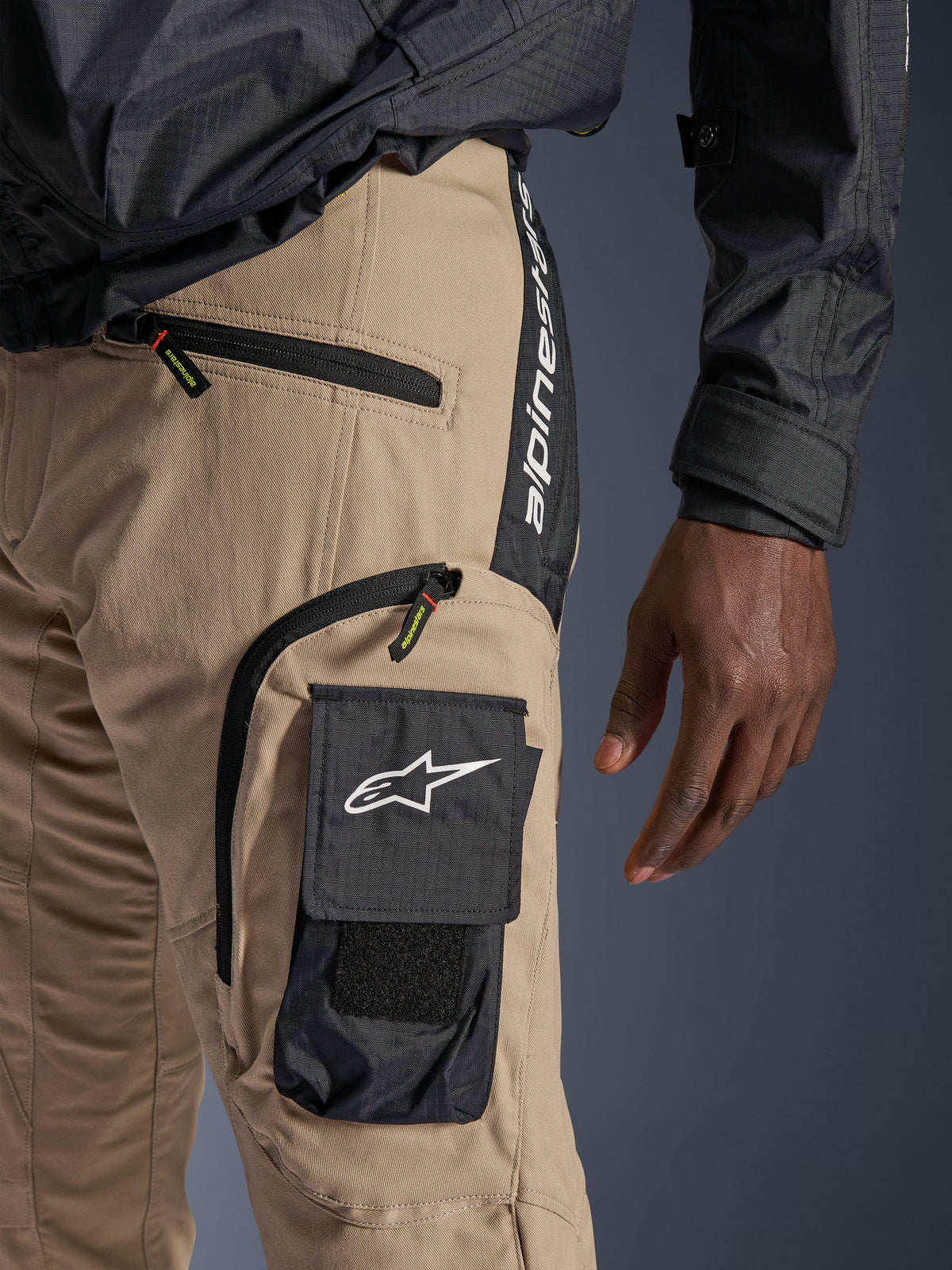 Pantalon Alpinestars Acteon Sable, vue détaillée de la hanche et de la cuisse, avec une poche cargo latérale noire ornée du logo étoile blanc, des poches zippées et le marquage Alpinestars vertical sur un empiècement noir, vêtement de moto urbain.