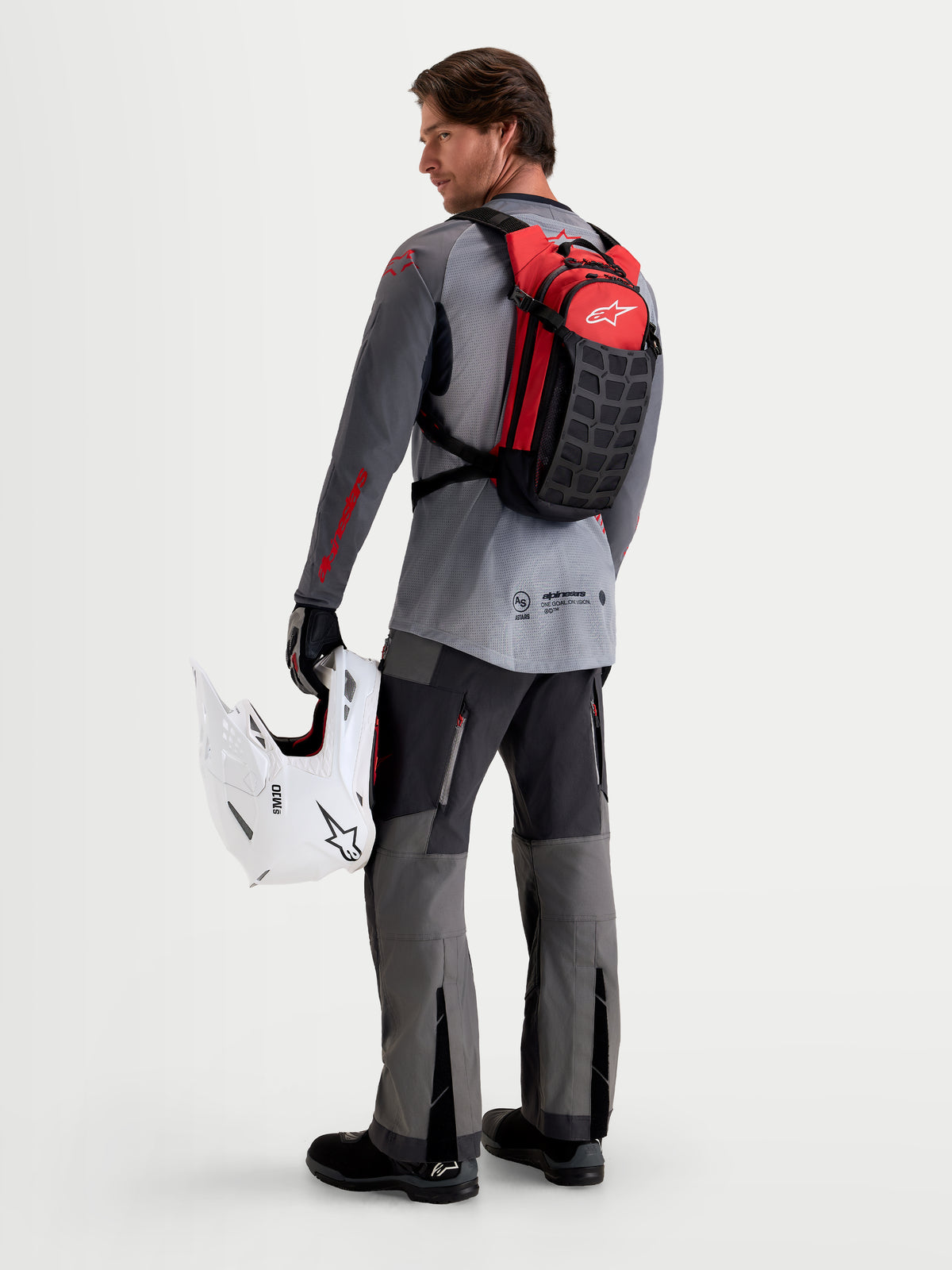 Alpinestars AT-9 Rucksack, Motocross-Rucksack in Schwarz und Leuchtrot, Rückansicht des Modells mit weißem MX-Helm und passender Ausrüstung, leichte Textilkonstruktion für den Einsatz auf der Rennstrecke und im Fahrerlager