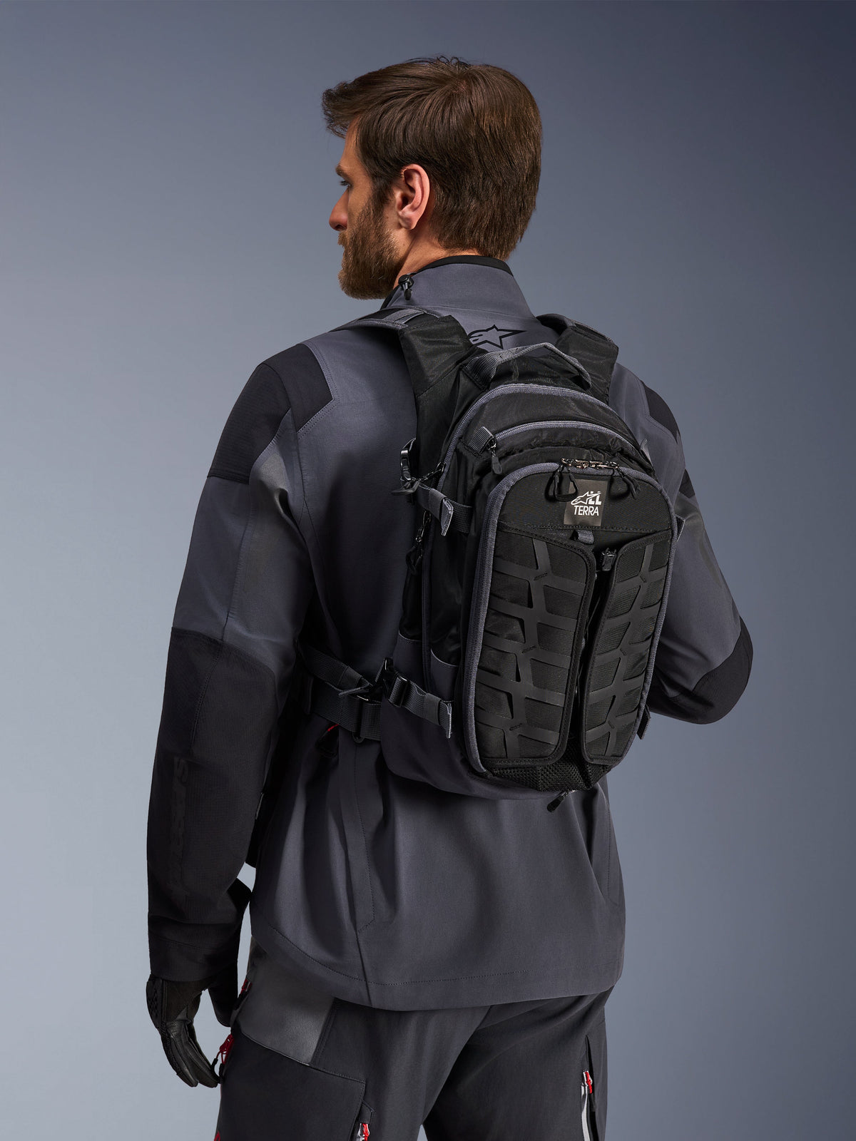 Alpinestars AT-15 rugzak, adventure motorrugzak, zwart en diepgrijs, gedragen door een rijder in een grijze jas, met een technisch ontwerp met MOLLE-webbing en 'TERRA'-branding, ergonomische off-road uitrusting