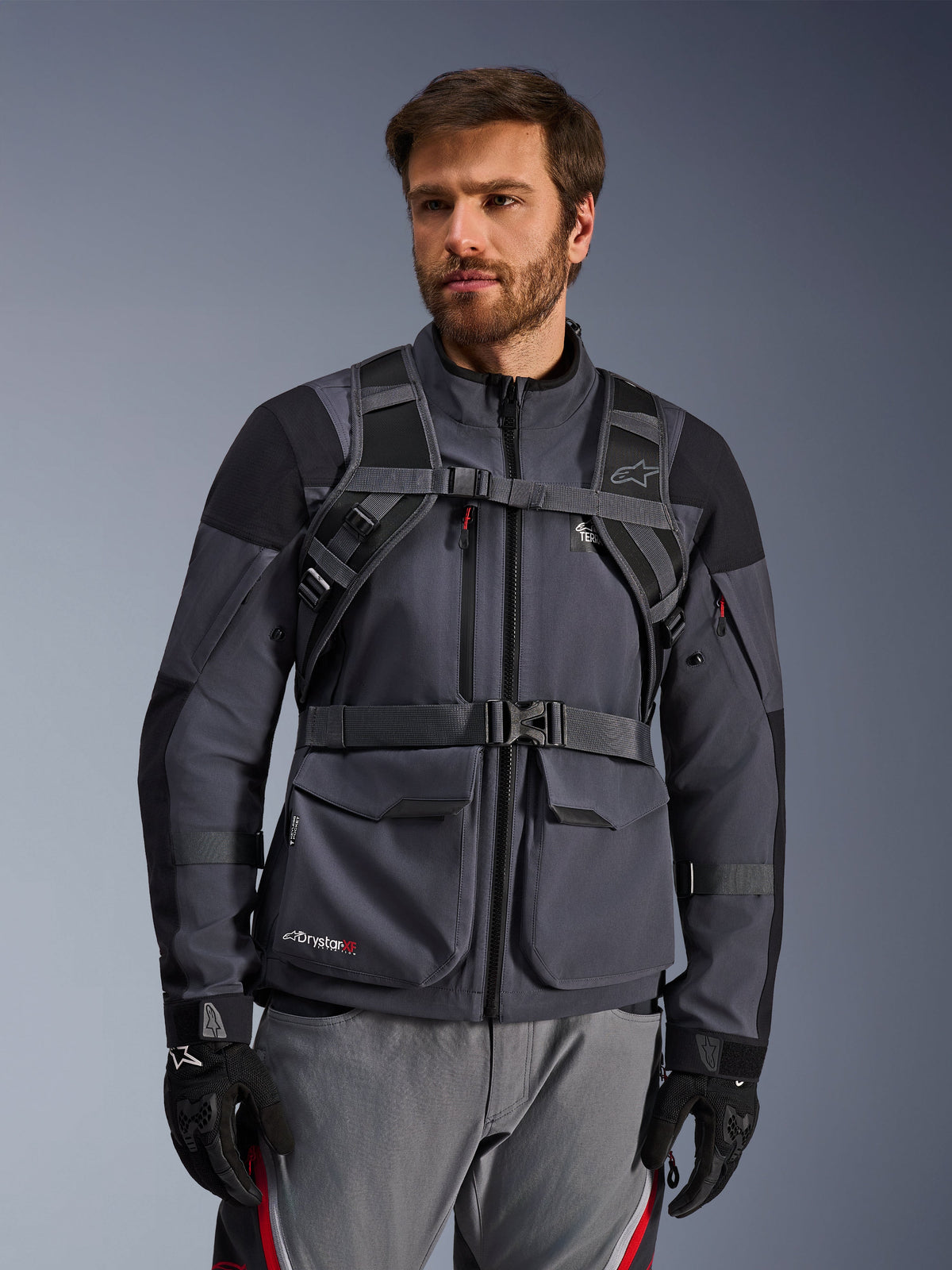 Alpinestars AT-15 rugzak draagsysteem, Black Deep Gray, vooraanzicht gedragen door een motorrijder over een grijze motorjas, voorzien van magnetische borstvergrendeling en stevige tailleriem, gecombineerd met zwarte handschoenen voor adventure motorrijden.
