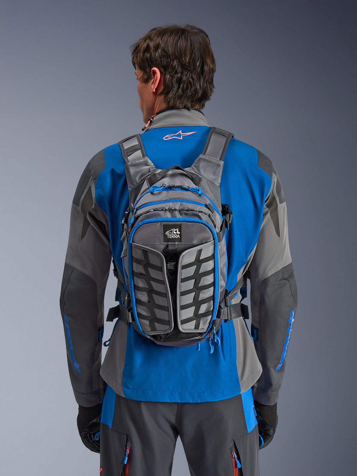 Alpinestars AT-15 Rucksack, Adventure-Motorradrucksack, Dunkelgrau und Tiefgrau, getragen von einem Fahrer über einer blau-grauen Jacke, mit All-Terra-Branding, ergonomischem strukturellem Design, verstellbaren Riemen und blauen Reißverschluss-Akzenten.