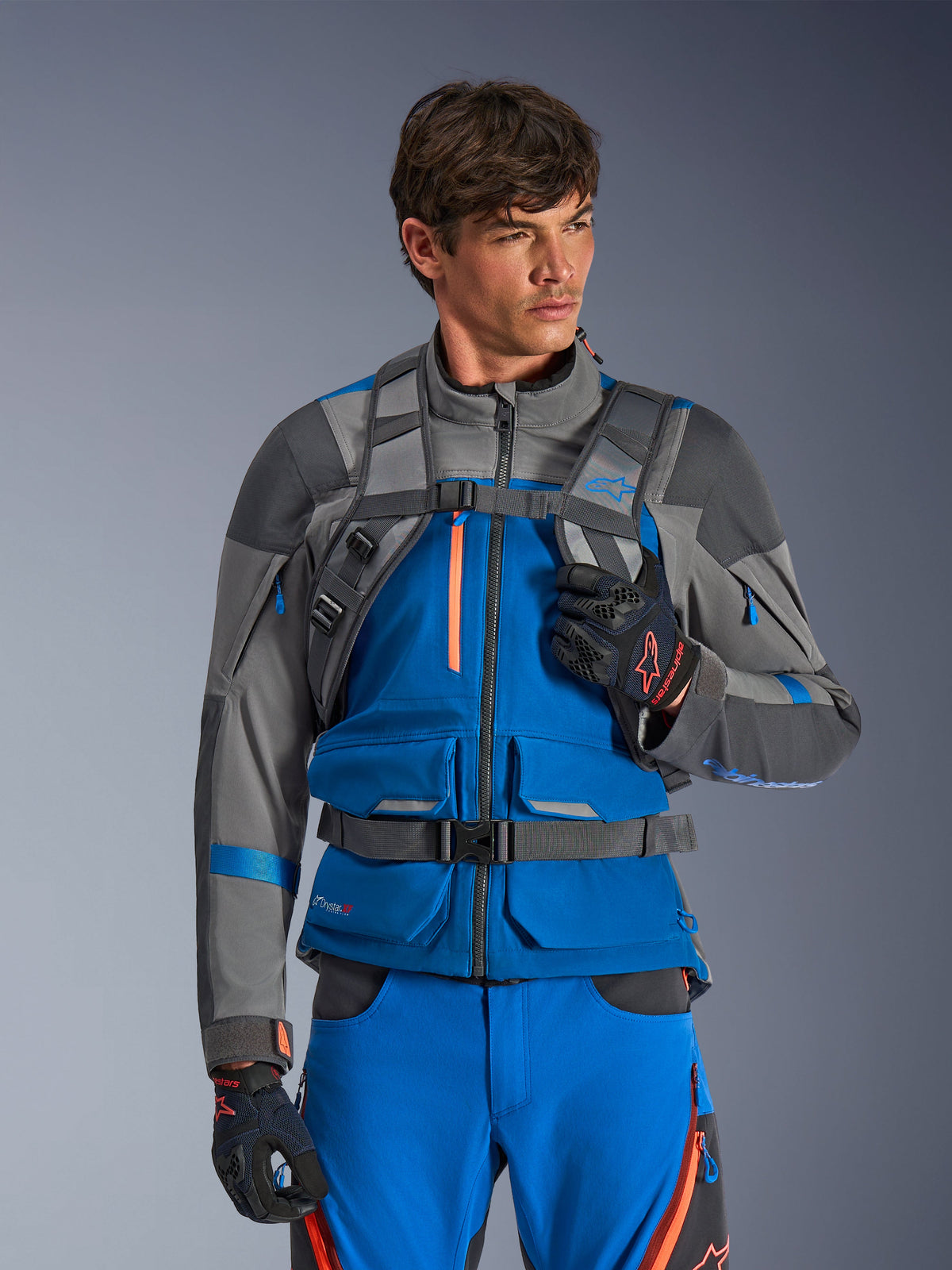 Alpinestars AT-15 Rucksack, Motorrad-Rucksack, Dunkelgrau, getragen von einem Fahrer in einer blau-grauen Adventure-Jacke und -Hose, mit technischen grauen Schultergurten, magnetischem Brustverschluss und Hüftgurt, robustes Design für Offroad-Fahrten