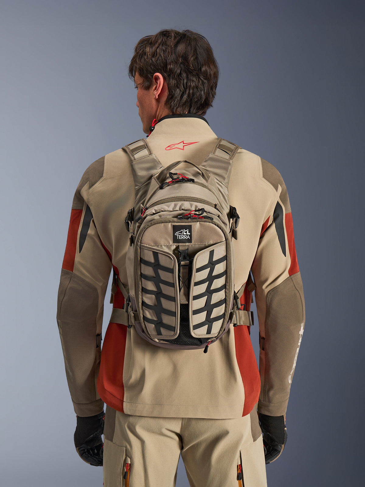 Alpinestars AT-15 rugzak, Aluminum Walnut, technische adventure motorrugzak gedragen door een rijder in een beige en rode jas, voorzien van bandenprofiel-patroon, TERRA-logo, rode ritslipjes en verstelbare banden