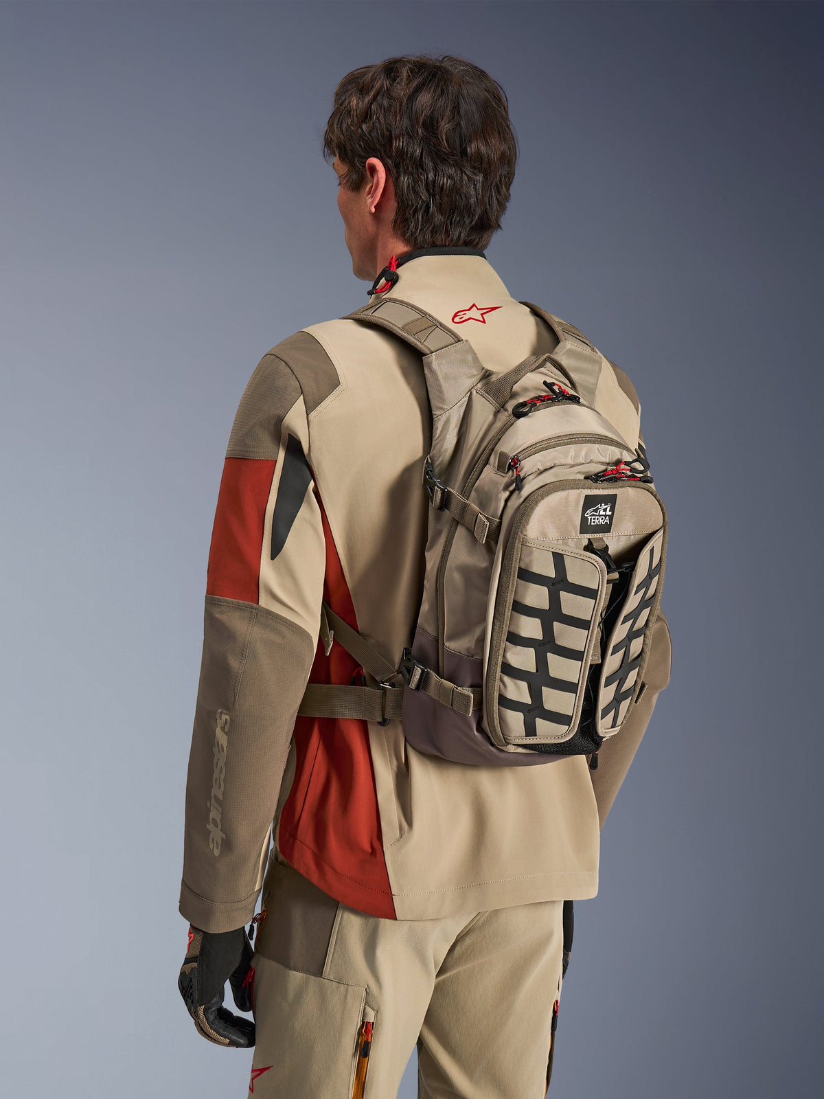 Alpinestars AT-15 Rugzak in Aluminum Walnut, technische motorrugzak gedragen door een rijder in bijpassende adventure-uitrusting, voorzien van een textielconstructie in tan en bruin met zwarte structurele accenten en rode ritslipjes, ontworpen voor off-road en adventure rijden.
